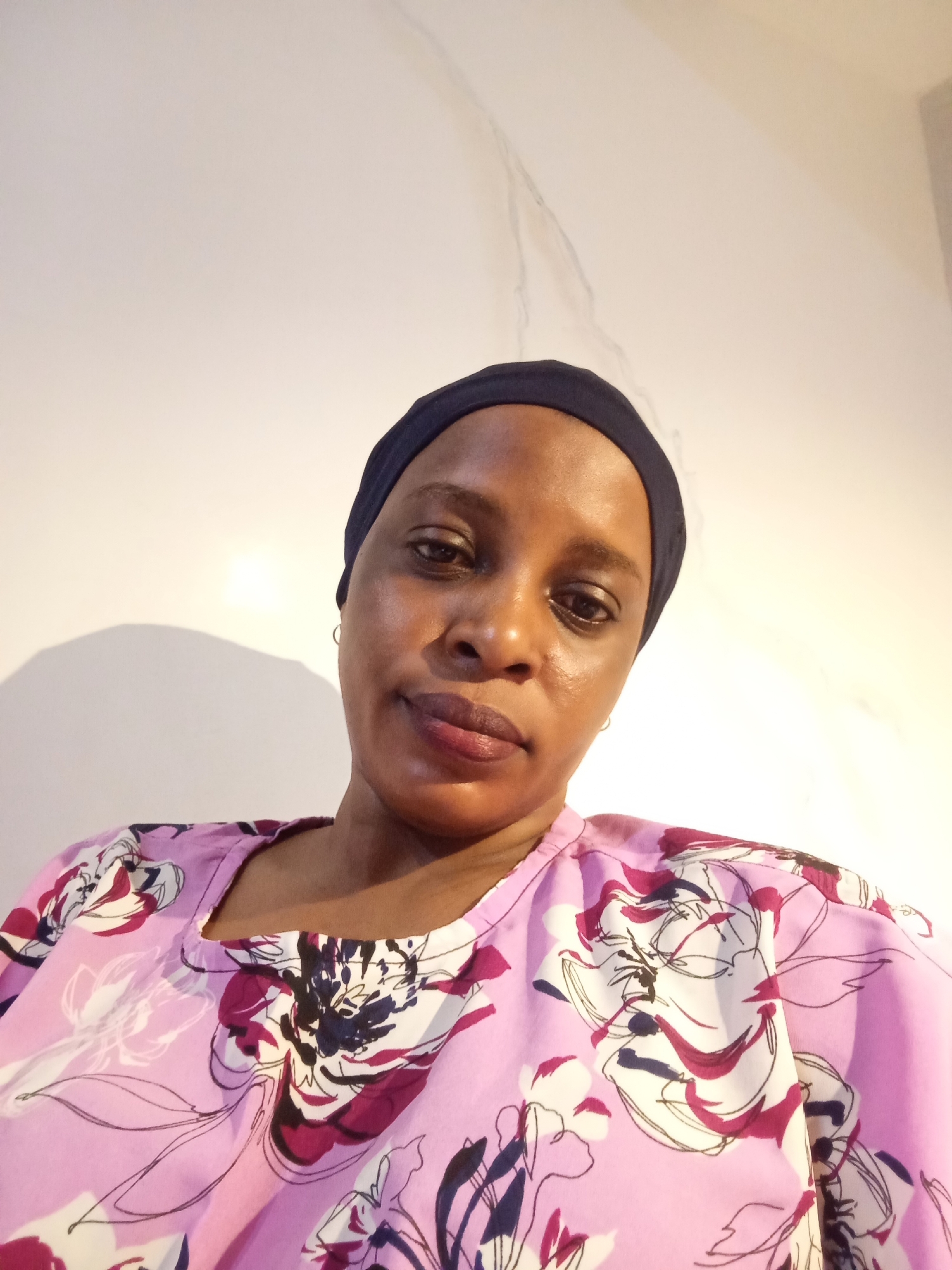 The image depicts a woman wearing a navy blue headwrap and a pink floral print top. The background is a plain white wall with a slight shadow. The subject appears to be in a room, possibly indoors. There is no clear indication of Unguwar Rimi, Nigeria. No specific actions or interactions are evident in the photo.