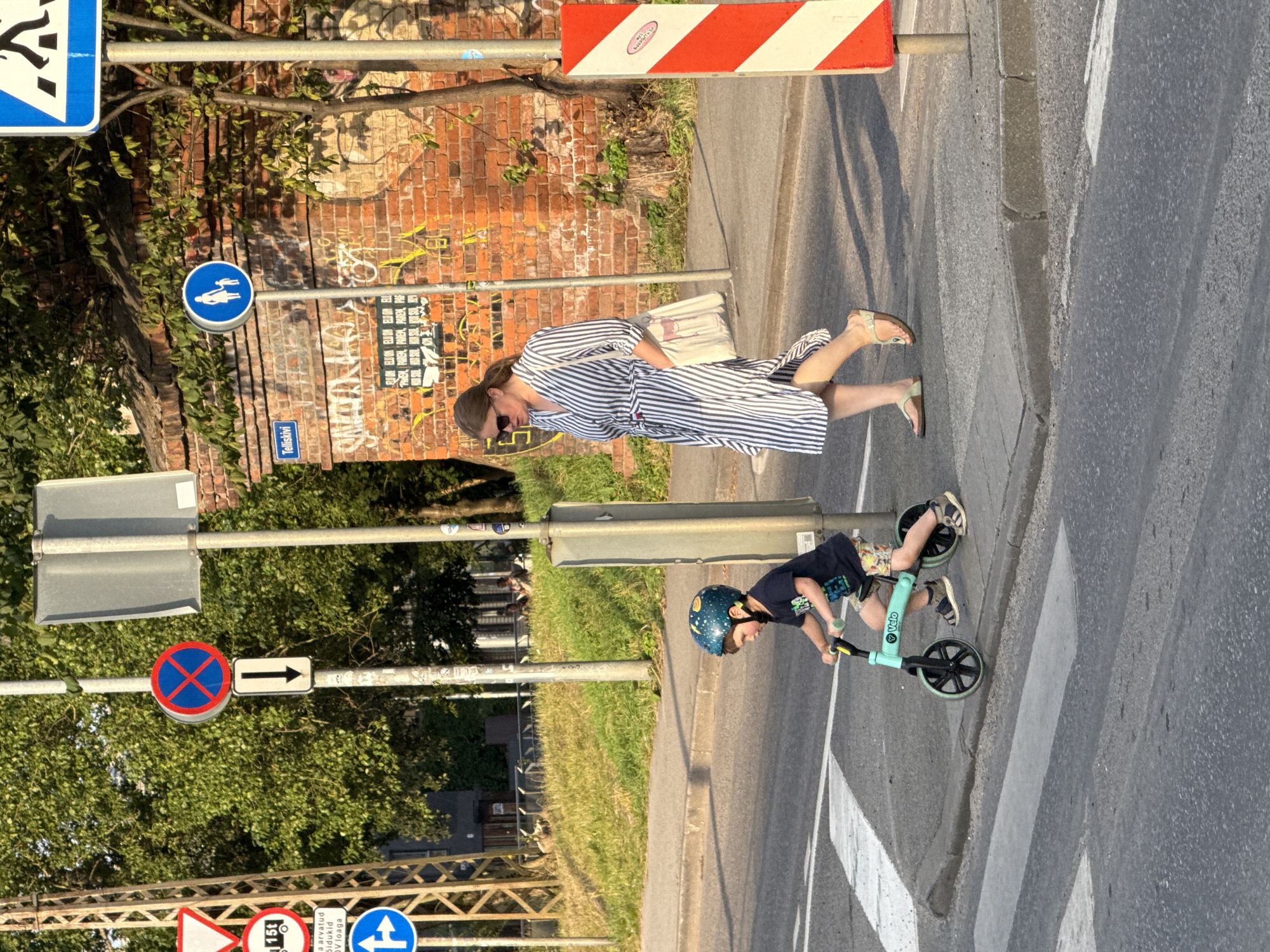 The image shows a street scene in Tallinn, Estonia, near the Telliskivi Creative City. A young boy wearing a helmet is riding a balance bike on a pedestrian crossing. He is wearing a dark blue t-shirt and patterned shorts. A woman, likely his mother, is walking behind him on the sidewalk. She is wearing a striped dress and sandals, and carrying a tote bag. The background includes a brick building with graffiti, trees, and traffic signs. The lighting suggests it is late afternoon or early evening.