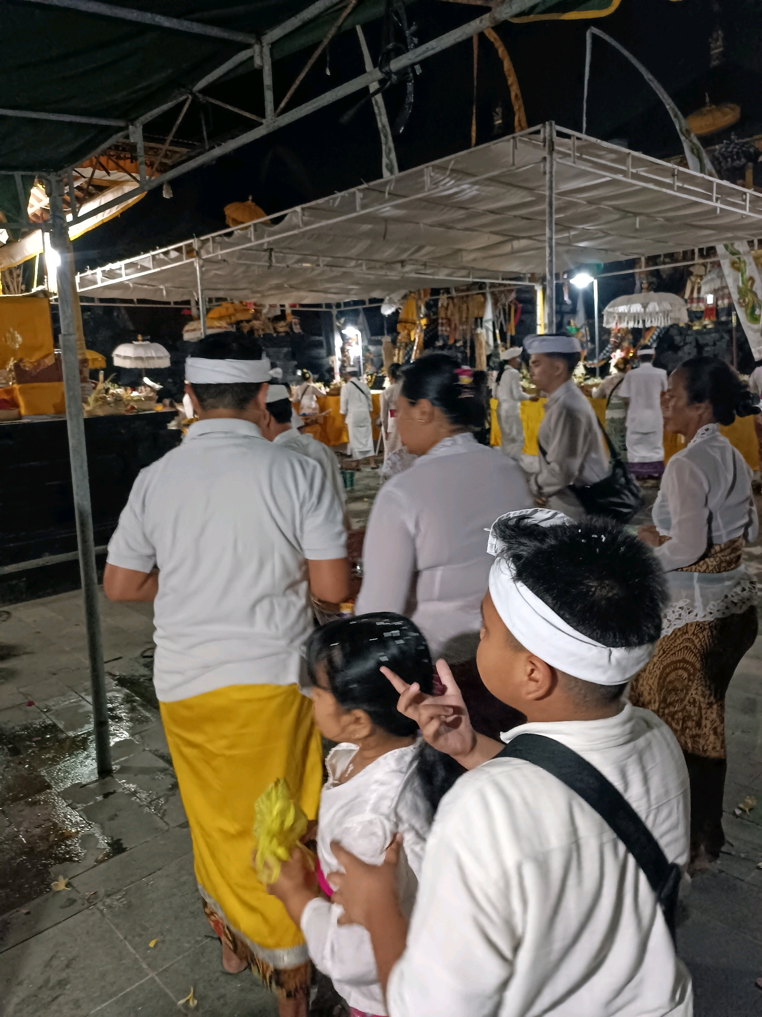 The image captures a nighttime scene at a temple or sacred site, possibly during a religious ceremony. Several individuals are present, many wearing traditional white clothing and headbands, which are common attire for Balinese ceremonies. In the foreground, a young boy with a white headband and a white shirt with a black strap across his chest is visible from the back. To his left, a young girl in a white outfit is also seen from the back, holding a yellow object, possibly a flower or offering, and making a peace sign with her fingers. Further back, other attendees are observed, including men in white shirts and yellow sashes and women in white blouses and patterned skirts. The background reveals a structure with a white tarp roof supported by metal poles, illuminated by artificial lighting, creating a festive or ceremonial atmosphere. Umbrellas and decorative elements are also visible. The ground appears to be paved with stone, and there are some wet patches, suggesting recent rain or cleaning. The overall ambiance is one of reverence and community gathering. Based on available information, this scene is consistent with the cultural and religious practices found in and around Kediri, Indonesia, which has a significant Hindu population and numerous temples where such events are held.