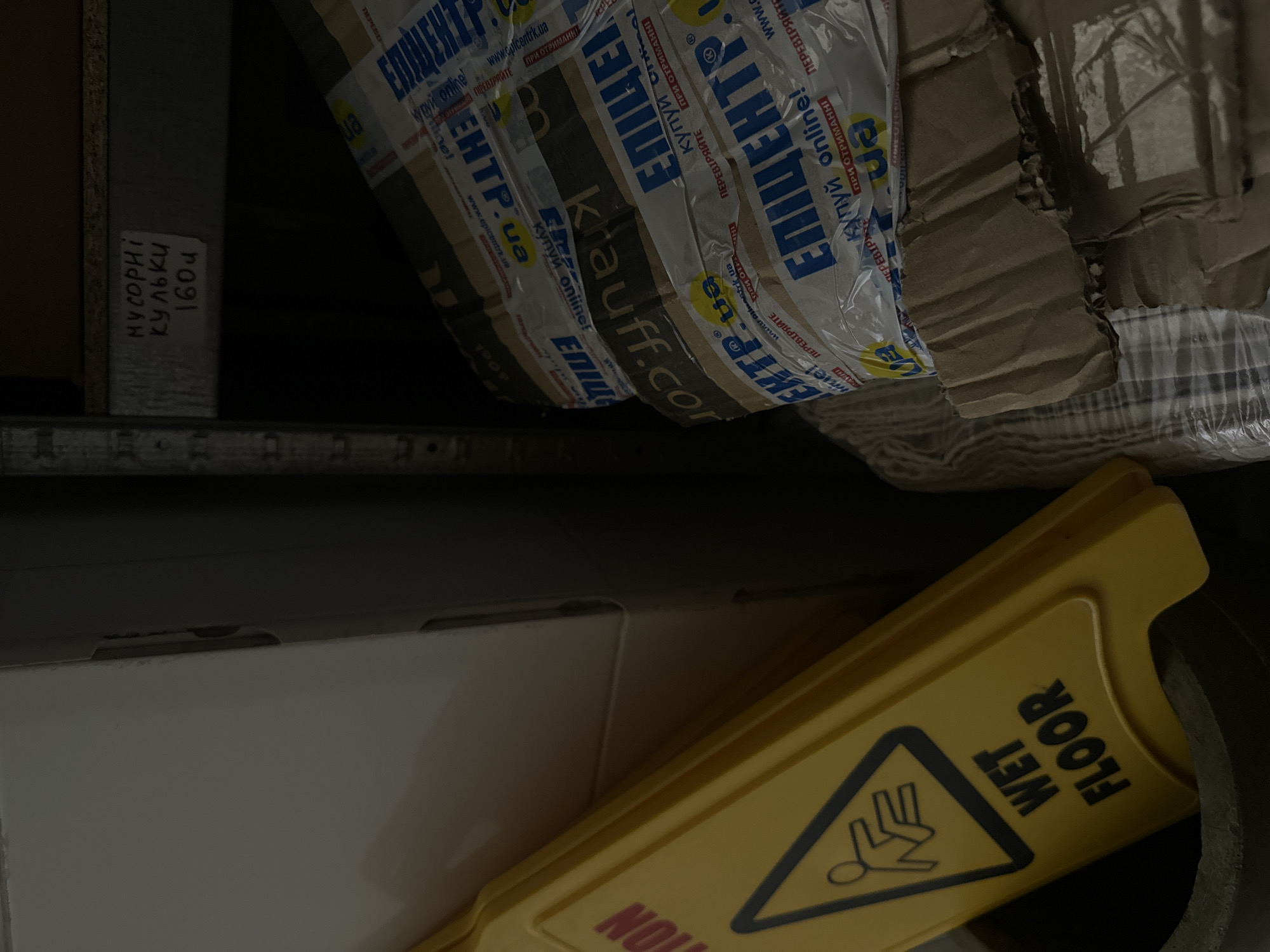 The image depicts a cluttered storage area, possibly in a warehouse or back room, located in Ternopil, Ukraine. A yellow "Wet Floor" sign is prominently displayed in the foreground, leaning against a white box. The sign features a black pictogram of a person falling. Behind the sign, several cardboard boxes are stacked and wrapped in clear plastic tape. The tape has visible branding, including Cyrillic text that appears to say "ЕПІЦЕНТР" and "KRAUFF.COM", suggesting they are shipping or product packaging from a retail or wholesale company. In the upper right corner of the image, a shelf is visible with a handwritten label in Cyrillic that reads "МУСОРНІ КУЛЬКИ 1604", which translates to "Trash bags 1604". The overall environment appears dim and disorganized, indicative of a storage space rather than a public area.