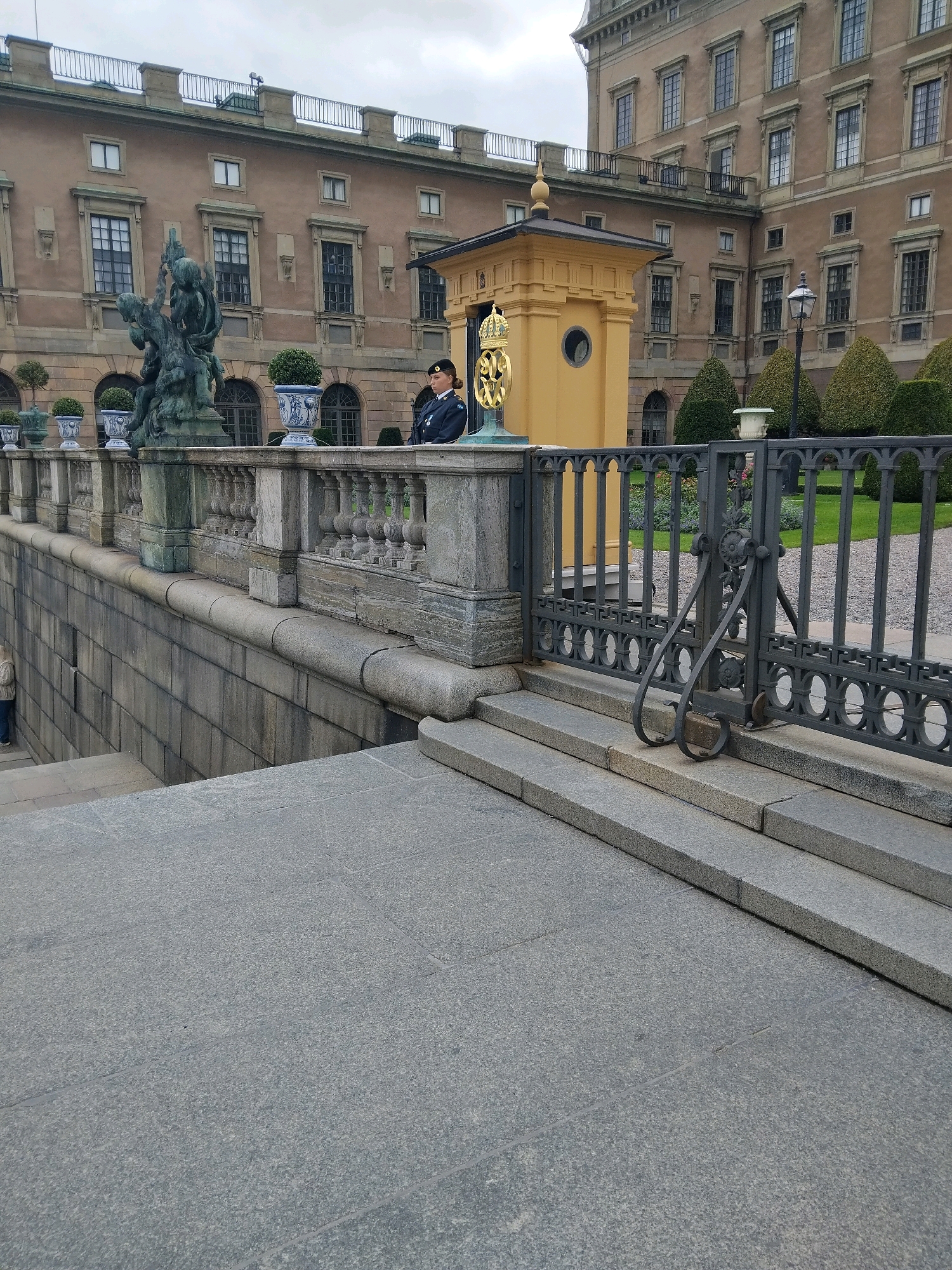 The image shows a view of the Royal Palace in Stockholm, Sweden. In the foreground, there is a grey stone staircase leading up to a decorative metal railing. Behind the railing, a soldier in a dark uniform stands at attention next to a small yellow guardhouse. The soldier is wearing a dark beret and a dark jacket with medals. The guardhouse is topped with a golden crown ornament. In the background, the large, tan-colored Royal Palace building is visible with its many windows. The sky is overcast. The scene depicts a formal setting, likely a ceremonial guard post.