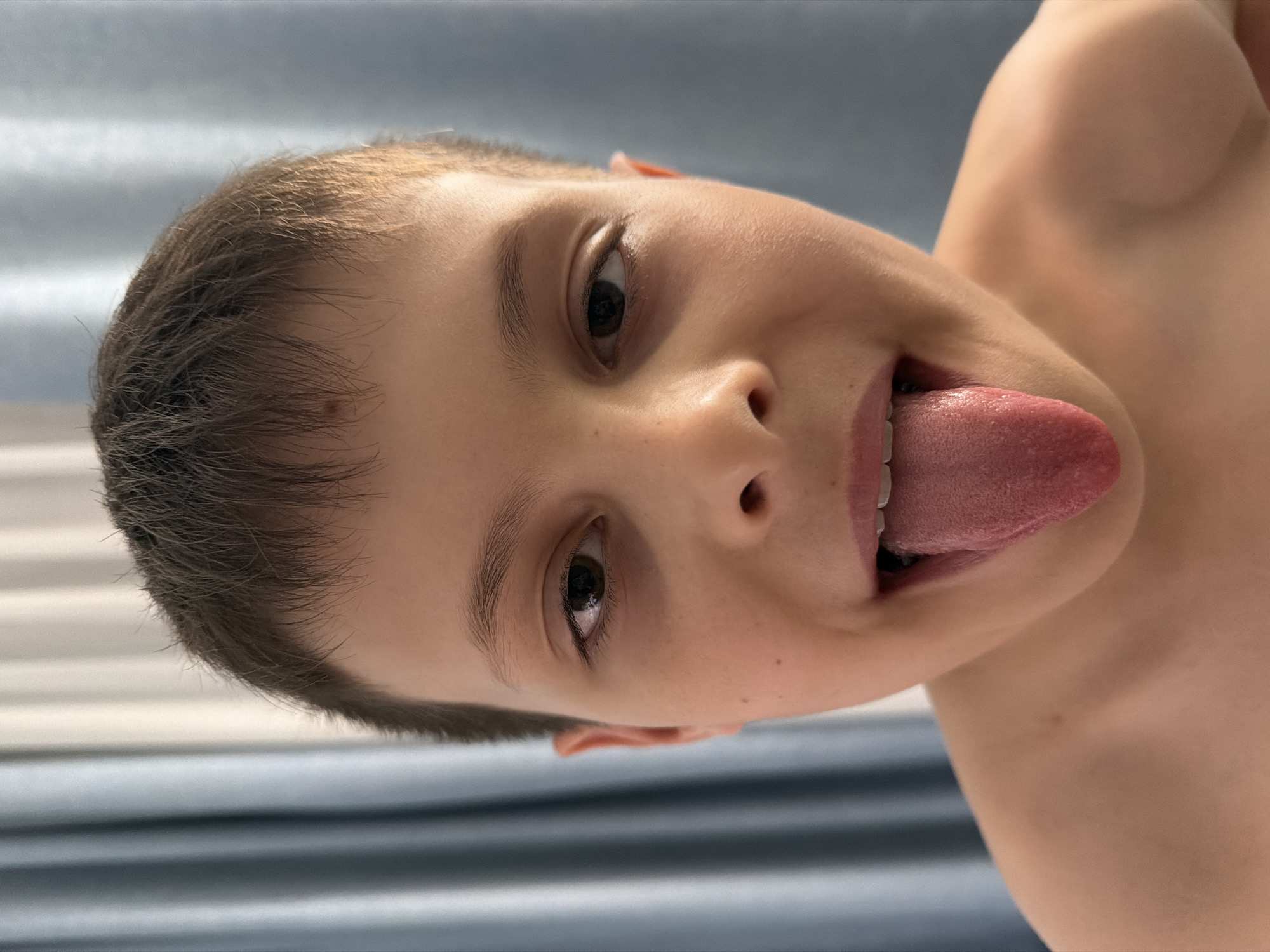 A close-up, tilted perspective shot depicts a fair-skinned young person with short brown hair. The person's mouth is open, and their tongue is extended outward. The individual has brown eyes and a visible mole on their face. The background appears to be a blurred blue curtain. It is not possible to determine the location in Votkinsk, Russia based on the image.