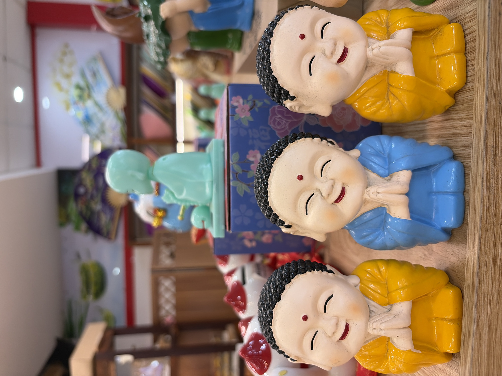 Three small, decorative figurines of young monks are positioned in the foreground of the image. The monk on the left and the monk on the right are wearing yellow robes, while the monk in the middle is wearing blue robes. All three figurines have closed eyes, smiling mouths, and their hands are clasped together in a prayer-like gesture. They are placed on a wooden surface. In the background, there are other decorative items and shelves, suggesting this is a display or retail setting. The overall scene appears to be indoors, with a blurred background that includes some decorative artwork on a wall. There is no discernible information to connect this image to the city of Đà Nẵng, Vietnam.