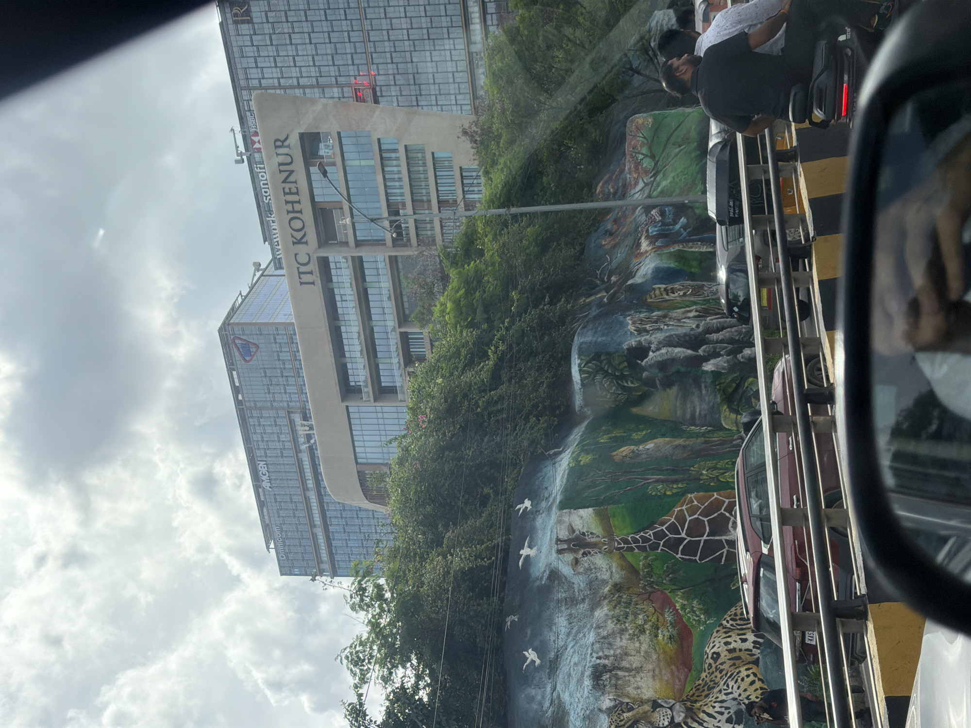 The image depicts a street view in Hyderabad, India, with a prominent mural on a rock face. The mural features various animals including a jaguar, a giraffe, and other wildlife, with birds depicted in flight above. In the background, modern high-rise buildings are visible, including the "ITC KOHE-SUR" building. The foreground shows a traffic scene with cars and a motorcycle with two riders. The sky is cloudy, suggesting overcast weather. The overall scene captures a blend of urban development and artistic expression within the city's environment.