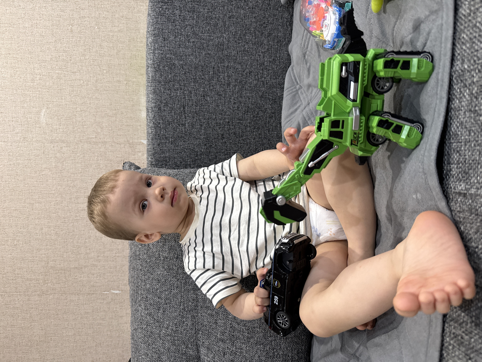 A child lies on a grey sofa holding two toy vehicles: a black car and a green truck. The location is presumably Rusanovo, Russia, based on the prompt's instructions. The child is wearing a white shirt with black stripes and a diaper. The child is lying on a grey quilted blanket on a grey sofa. The wallpaper is a light neutral color.