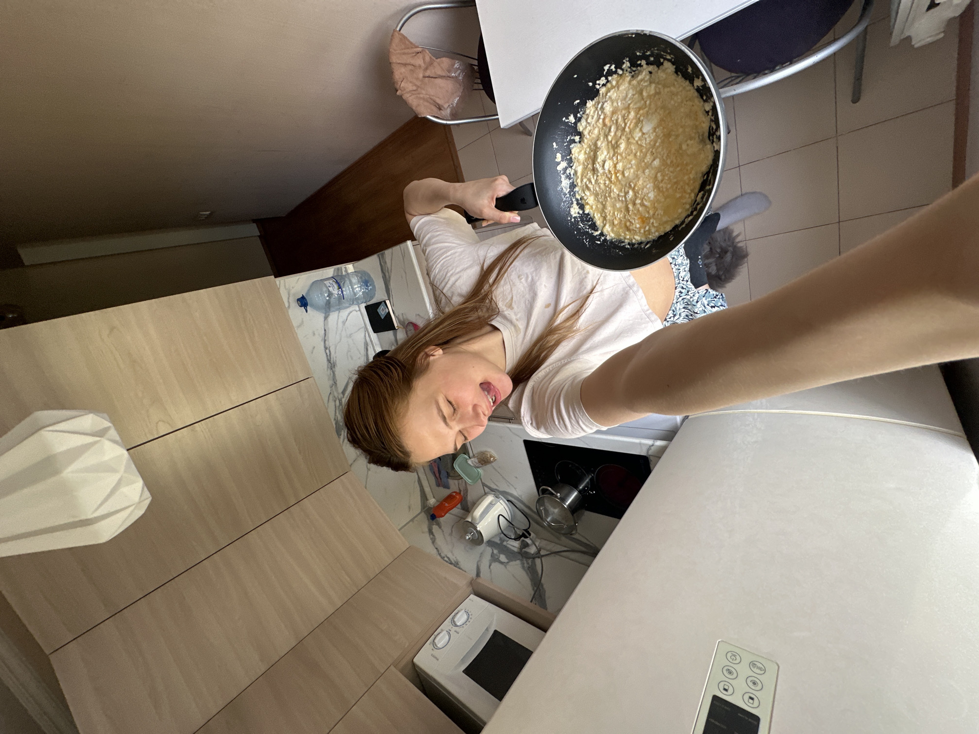 A person is taking a selfie in a kitchen. The person, who appears to be female with long hair, is holding a frying pan with cooked food in it. The kitchen has light wood cabinets, a marble-patterned countertop, a microwave, and a white refrigerator/oven unit. A dining table with chairs is visible. A water bottle is on the countertop. The exact location in Krasnoyarsk, Russia, cannot be determined from the image.