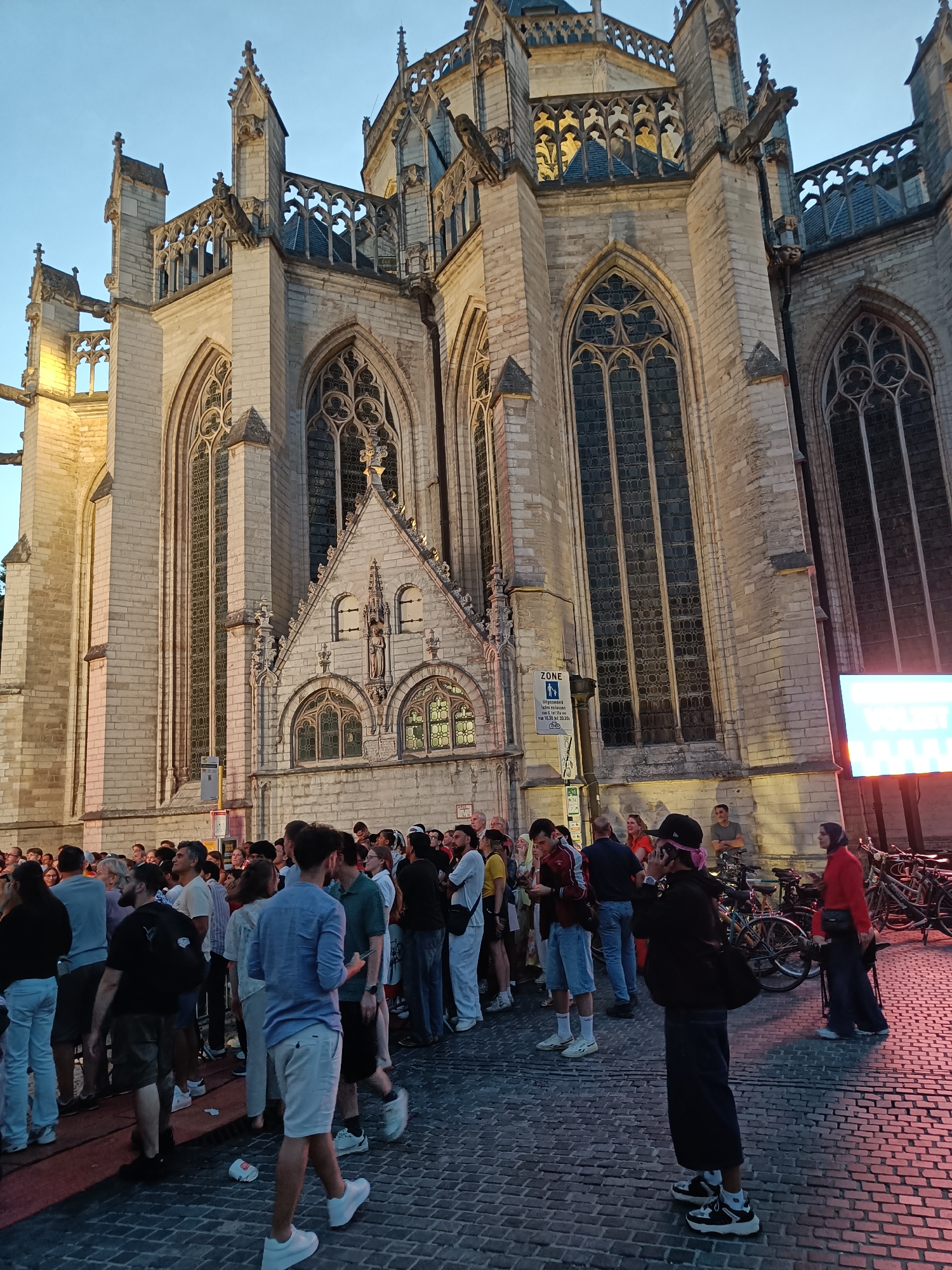 In the late evening in Leuven, Belgium, a crowd gathers in front of Saint Peter's Church. The imposing Gothic facade of the church, constructed from pale stone, features ornate architectural details, including flying buttresses, pointed arches, and large, intricate stained-glass windows. Gargoyles perch on the upper levels, and decorative stonework adorns the building.
The ground is paved with cobblestones, illuminated by the warm glow of streetlights and a large, brightly lit screen on the right side of the frame, displaying white text. A diverse group of people, dressed in casual attire, stand or mill about. Some are engaged in conversation, others are looking at their phones, and a few appear to be taking pictures. One individual in the foreground is wearing a black baseball cap backwards and holding a pink bandana. Bicycles are parked near the right edge of the image, suggesting a mode of transportation common in the city. The sky is a deep blue, indicating twilight. A sign with the word "ZONE" and text in Dutch is visible on a pole. The overall atmosphere suggests a public gathering or event taking place in the heart of the historic city.