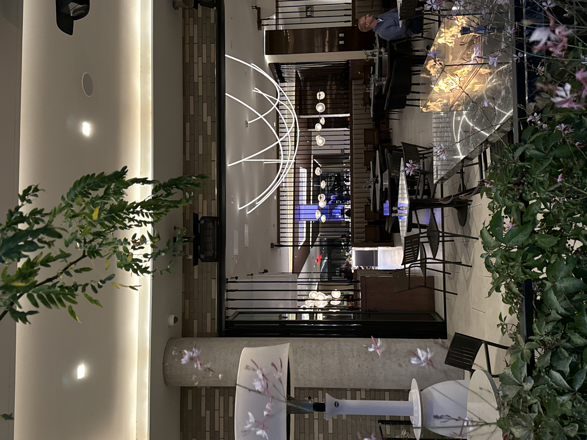 The image depicts an outdoor seating area of a restaurant at dusk. The restaurant appears to be located in a modern building with a brick facade. The seating area is furnished with dark chairs and tables, some of which have marble tops. A modern gas fireplace is visible on one of the tables, emitting warm flames. In the background, the interior of the restaurant is visible, with more tables and seating arrangements. A man wearing a blue shirt is seated at one of the tables, looking to the left. There is a large, white outdoor heater to the left of the frame, and a pillar with a floral projection. Lush green foliage with small pink flowers is in the foreground. The overall atmosphere is sophisticated and inviting.
The image is not located in Vancouver, United States of America.