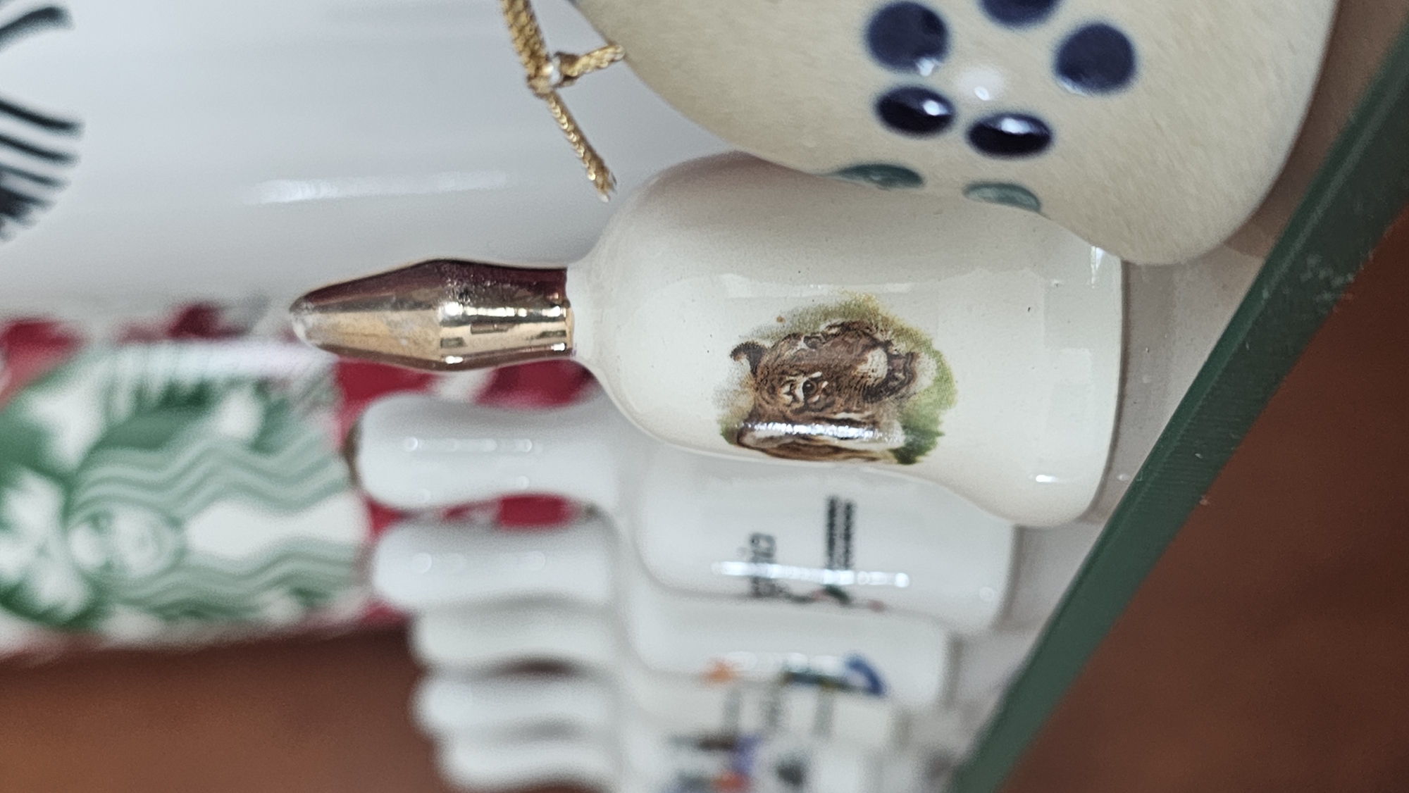 A close-up shot depicts a collection of small, white ceramic bells arranged on a surface covered in a green glass frame. The bells feature various painted designs, including a dog and other images. The background is blurred. The location, San Bernardino Tlaxcalancingo, Mexico, cannot be verified from the image alone.