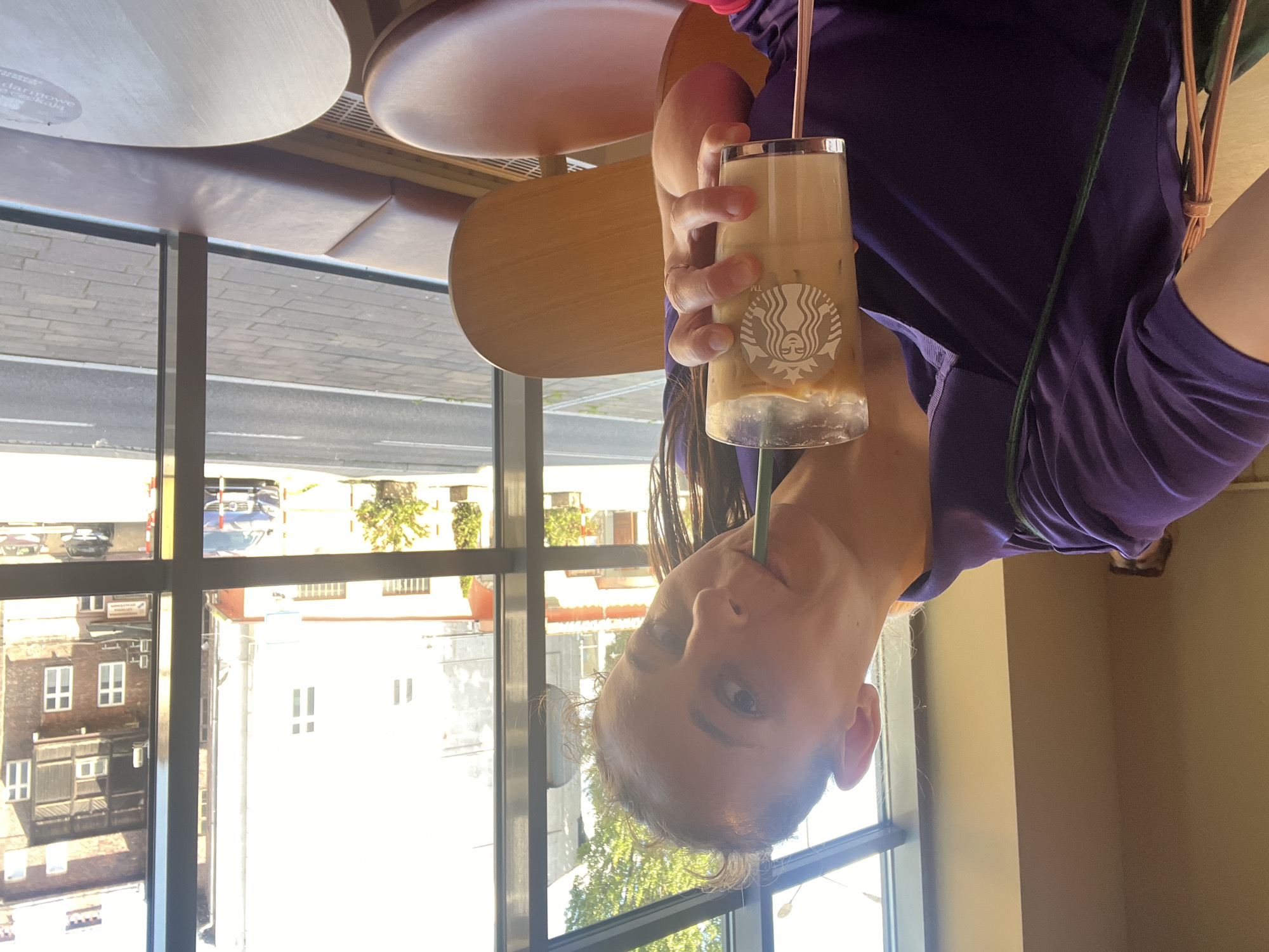 A woman in a purple polo shirt is holding a tall glass of iced coffee with the Starbucks logo. She is sipping the drink through a green straw. The woman has her hair pulled back and is looking at the camera. She is sitting in a cafe with large windows that look out onto a street. The cafe has wooden chairs and tables, and a padded bench along the window. Outside, there are buildings and cars.