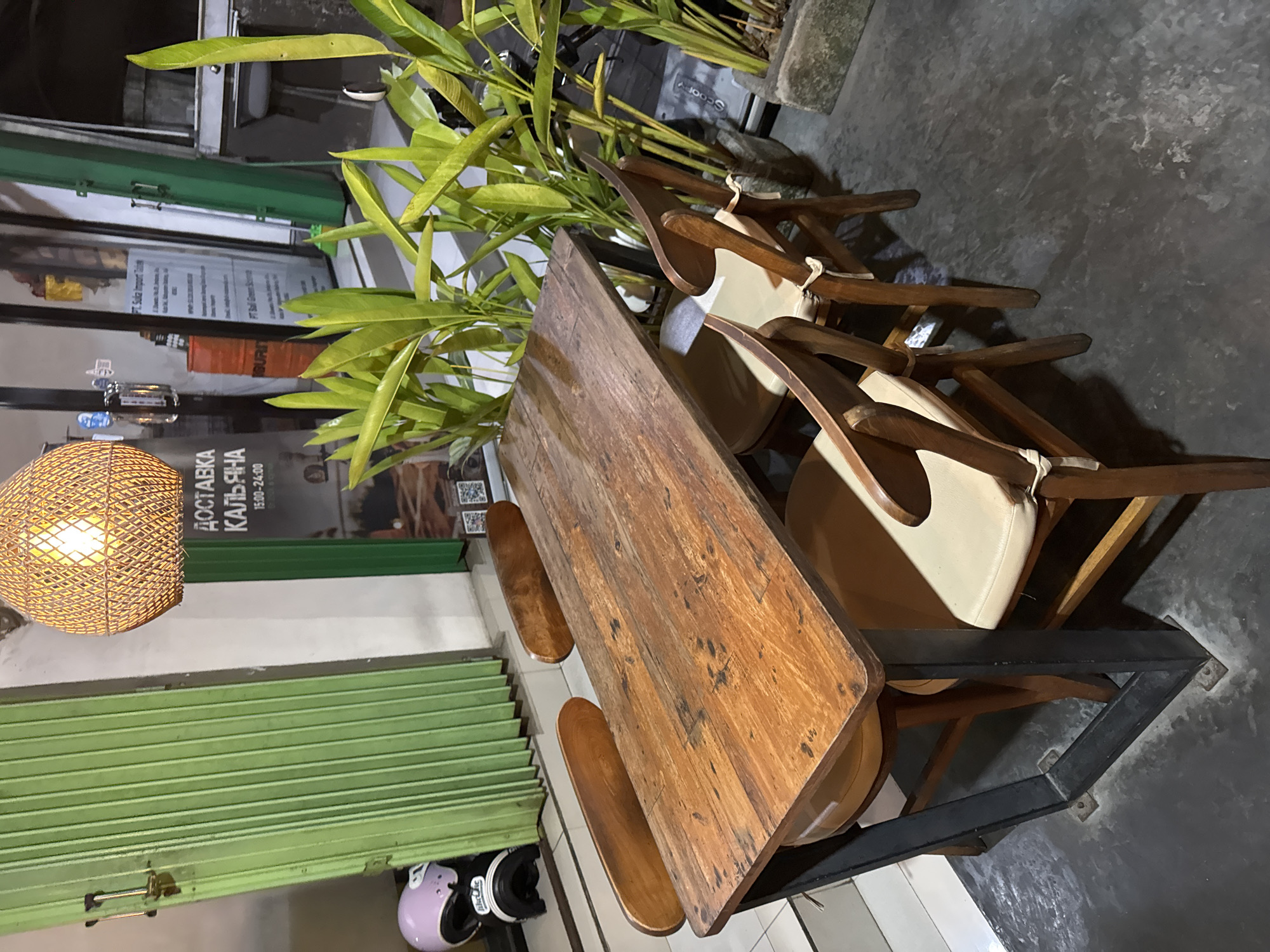 A wooden table with four chairs is set up outdoors in front of a green corrugated metal door. A string of lights hangs above the table. On the left, a pink and black motorcycle helmet rests on the ground. Plants are arranged in pots to the right of the table. The scene appears to be in Kuta Selatan, Indonesia.