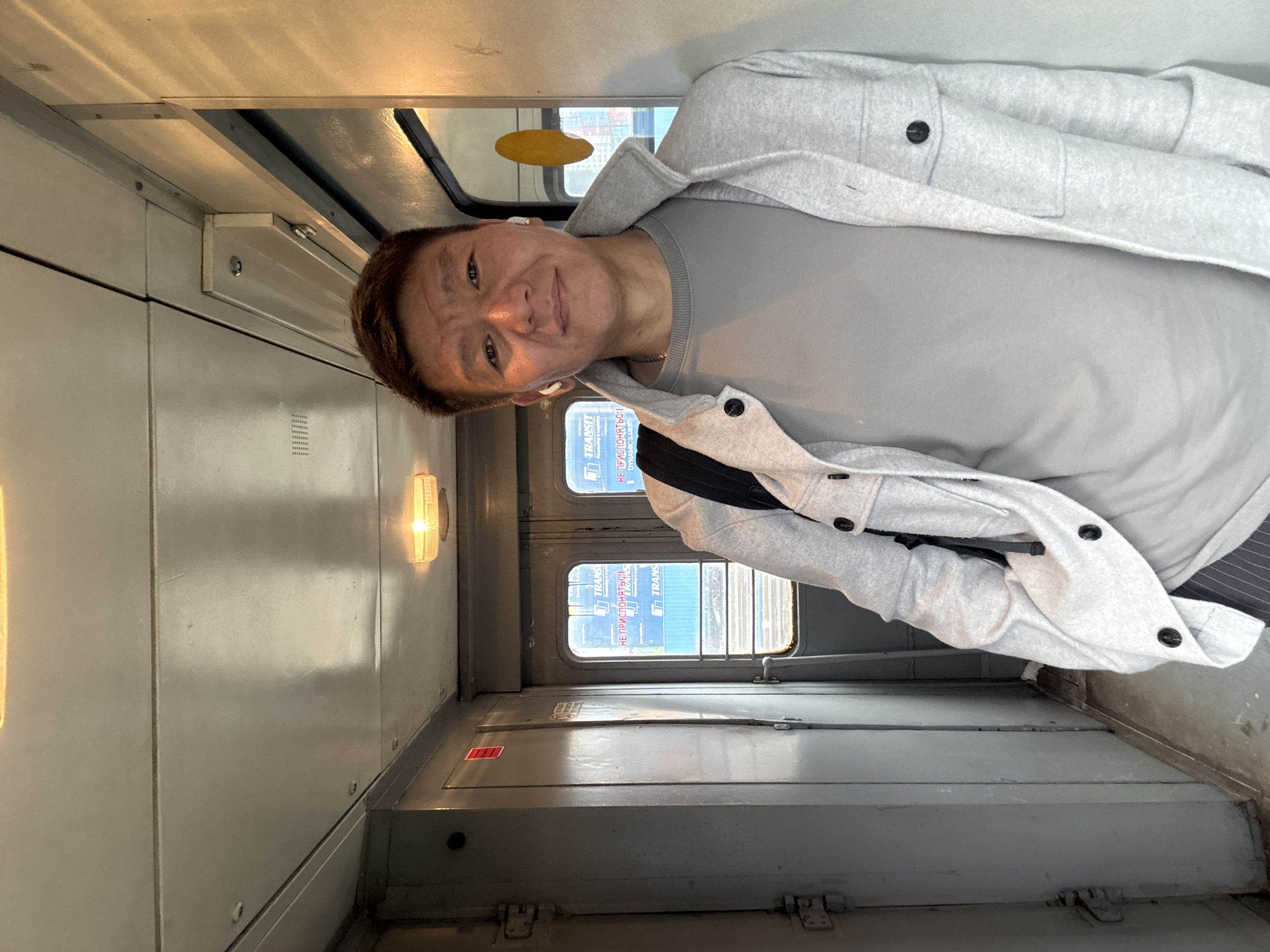 A medium shot captures a man standing in a narrow interior corridor of what appears to be a train car. He is facing the camera, with his body angled slightly to the left. The man is wearing a grey crewneck sweatshirt under a light grey jacket with black buttons and two chest pockets. He has dark hair, brown eyes, and is wearing white wireless earbuds. A black backpack is slung over his left shoulder.
The corridor has grey, paneled walls and a light-colored ceiling with two visible light fixtures. To the man's left, there are two closed doors with red stickers and visible hinges. To his right, there is a window with a view of an outdoor scene featuring blue signage with white text, indicating a "TRANSIT" company, and railway tracks. The ground is a concrete or tiled floor. The overall environment suggests a public transportation setting, possibly within Moscow, Russia, given the Cyrillic text on the visible signage.