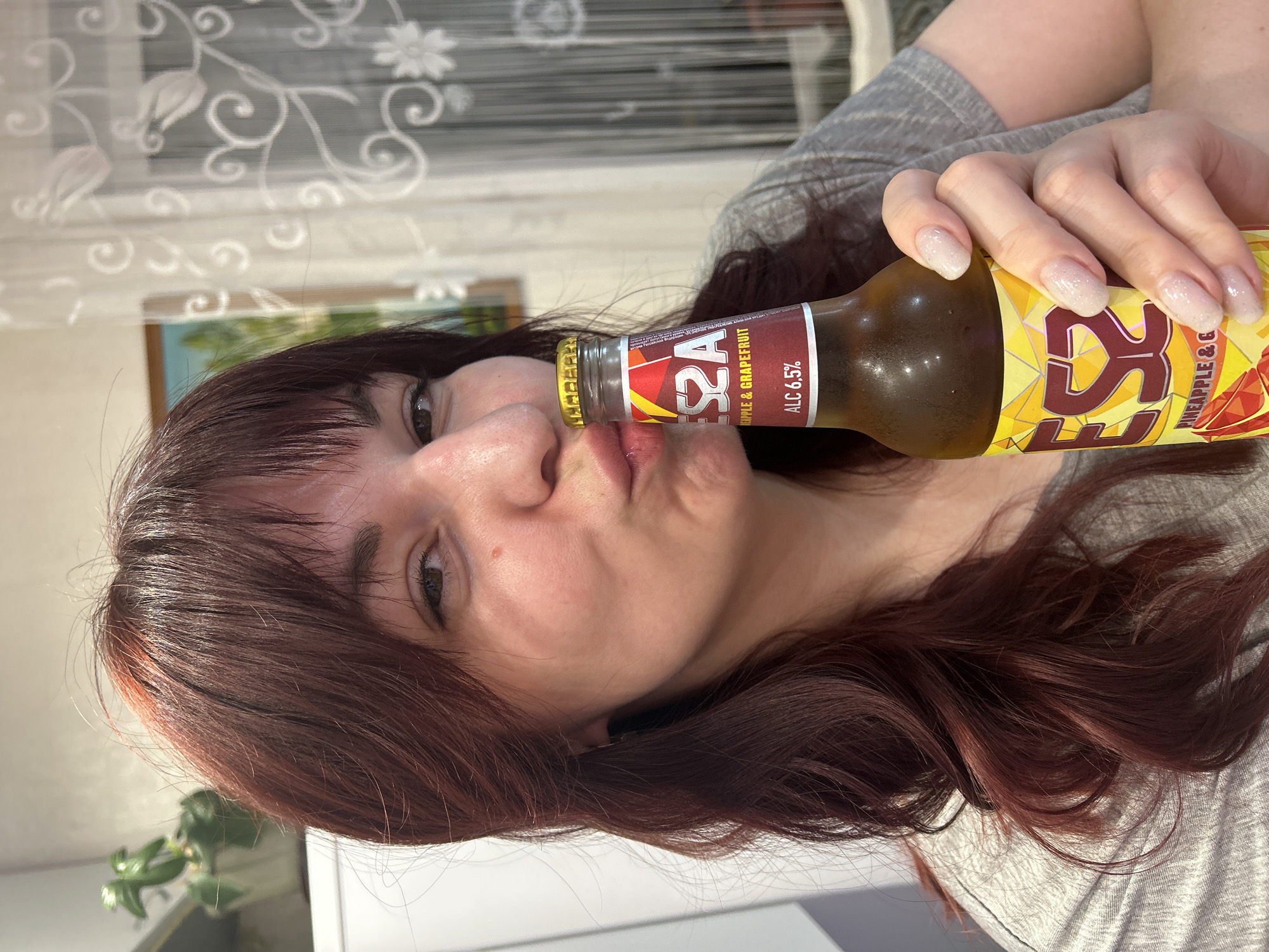 A woman with dark reddish-brown hair is holding a bottle of S2A Pineapple & Grapefruit beer in front of her lips. She has light skin and long, sparkly fingernails. She is wearing a gray shirt. The background includes a window with sheer curtains and what appears to be a houseplant. The location, Фоки, Russia, is not discernible from the image.