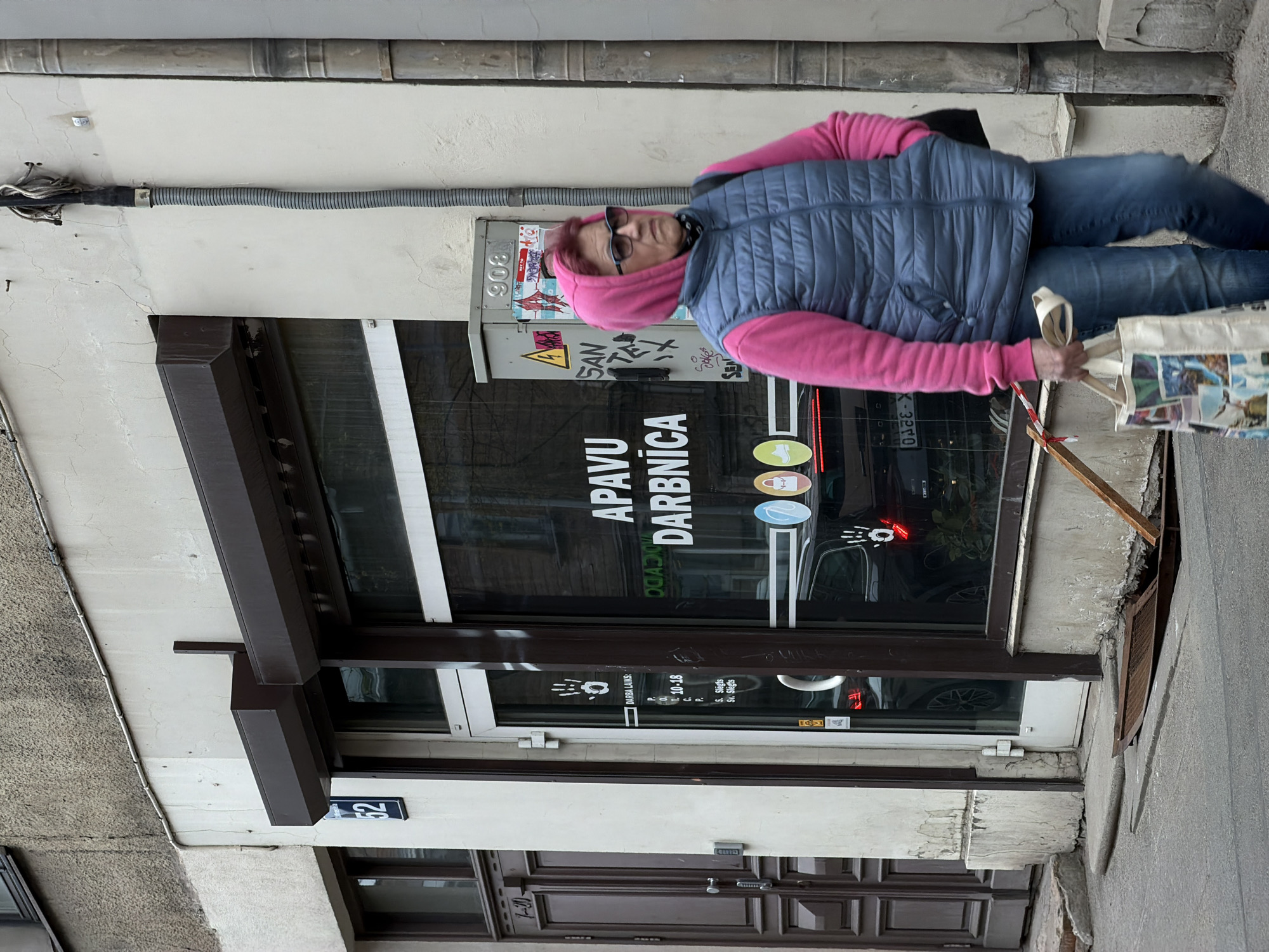 A horizontally oriented outdoor shot features a woman in a pink hoodie and blue vest carrying a tote bag and what appears to be a wooden frame. She is walking near a building that houses a shop called "APAVU DARBNĪCA" (Shoe Workshop), with an address number "52" visible on the wall. Graffiti is present on an electrical box next to the shop's window. The shop's business hours are posted as 10-18, with closed hours on Saturdays (S.) and Sundays (Sv.). The building's façade shows signs of wear and tear. The image suggests a location in Riga, Latvia, based on the Latvian language signage. The focus is sharp, and the scene is well-lit, providing clear visibility of the details.