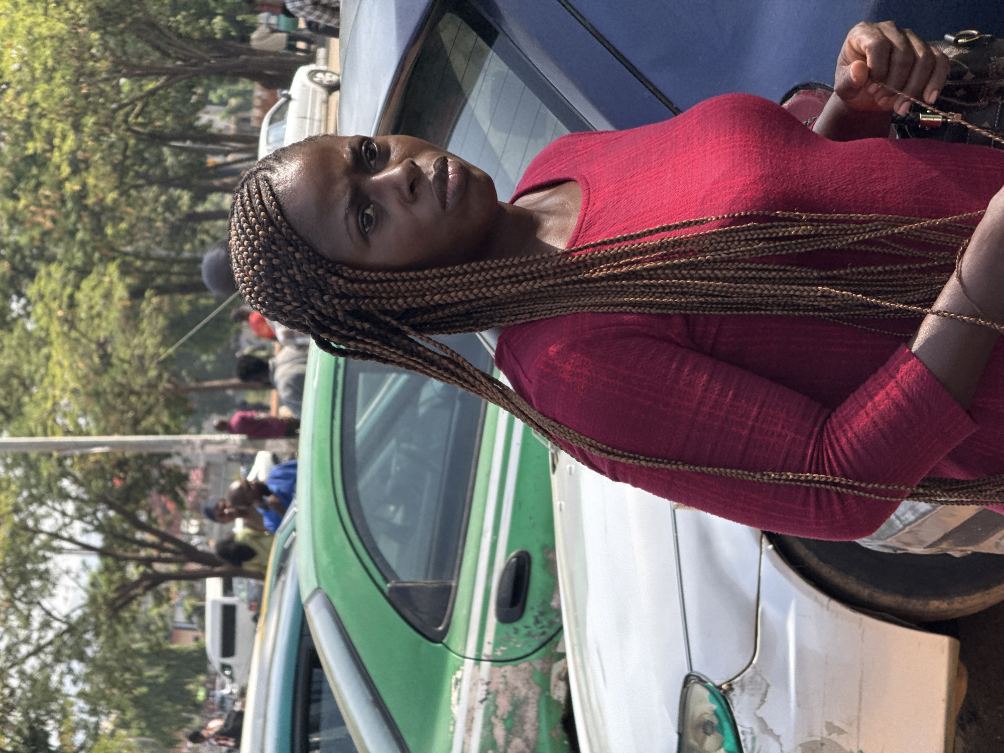 A medium-shot, portrait-oriented image captures a woman with dark skin and long, light brown box braids standing prominently in the foreground. She is wearing a reddish-pink long-sleeved top and is looking directly at the camera with a neutral to slightly serious expression. Her braids fall over her shoulders and chest. She holds a small, dark handbag with gold-colored hardware in her right hand, and a gold-colored bracelet is visible on her left wrist.
The woman is positioned next to a dark blue car, partially visible behind her. To her left, a green car with visible wear and tear is parked, and further to the right, a white car is partially visible. The scene unfolds outdoors during bright daylight, suggesting a sunny day with clear skies.
In the background, a street or road is lined with lush green trees, indicating a mix of urban and natural elements. Several blurred figures, presumably other people, are visible in the background, suggesting ongoing pedestrian activity. The overall setting is consistent with a public street in a city. No visible text is present in the image. The environment and the appearance of the vehicles align with the provided context of Abuja, Nigeria.