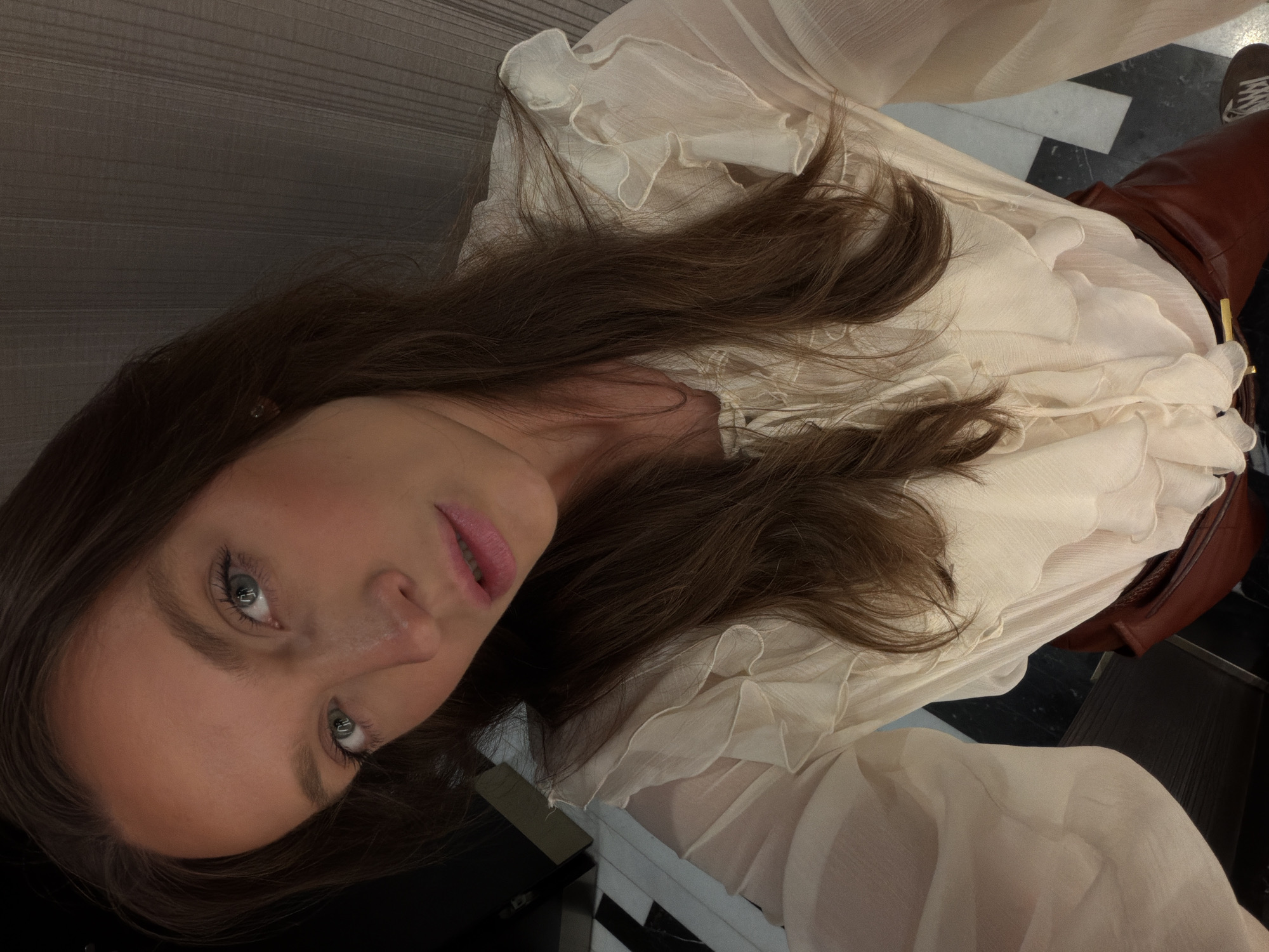 A woman with blue eyes and dark brown hair is posing for a photo. She is wearing a cream-colored, long-sleeved blouse with ruffles. Part of her lower body is also visible, showing brown leather pants and a brown shoe. The background is a mix of black and white checkered tile flooring and a gray wall. It is not possible to confirm the location of this image is in Budva, Montenegro.