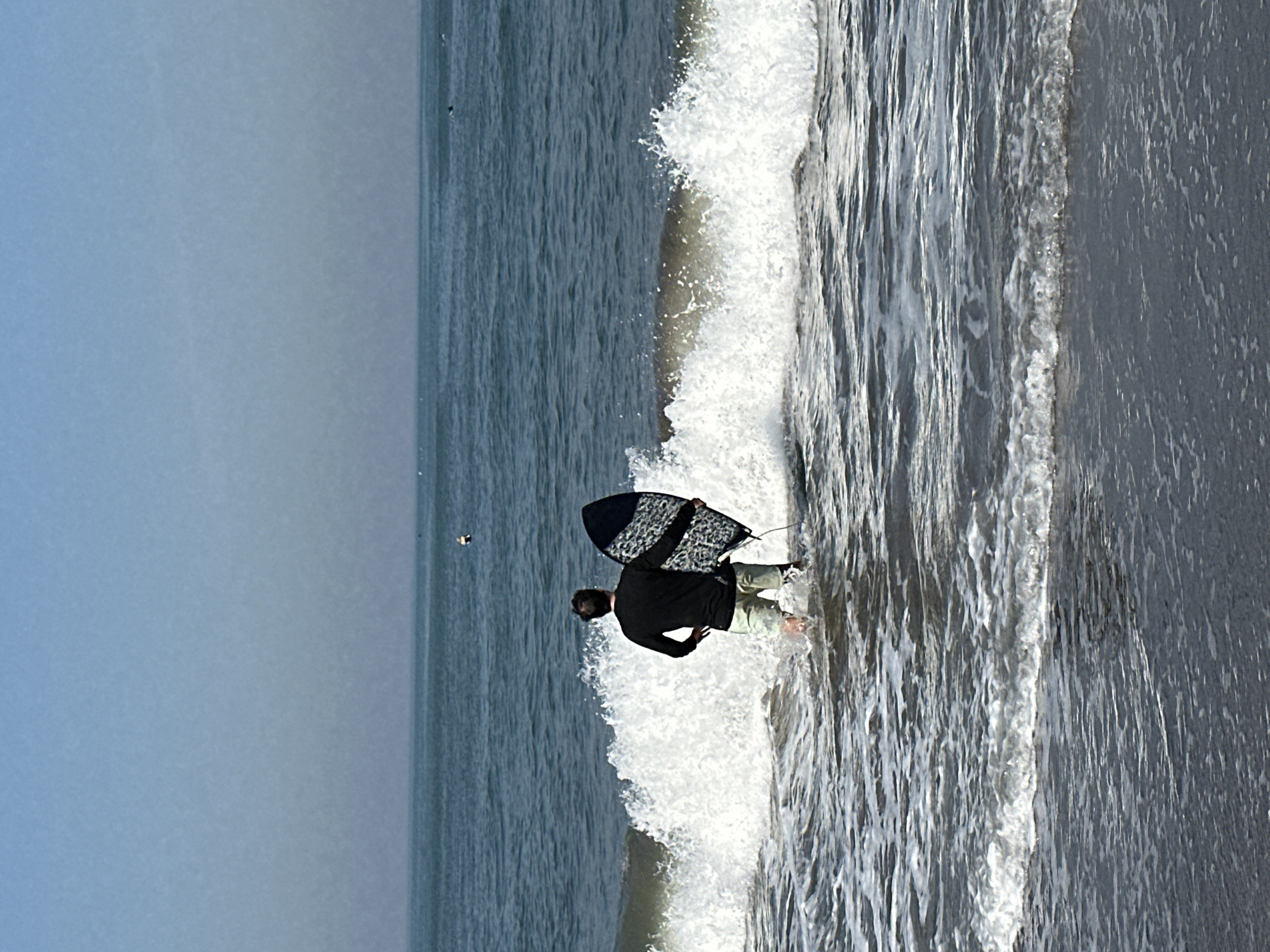 A person stands in shallow ocean water holding a skimboard. The person is facing away from the camera and is wearing a black long-sleeved shirt and light-colored shorts. The skimboard is black with a white and grey patterned design. White waves are breaking around the person's legs. In the background, the ocean extends to the horizon under a clear, hazy sky. The scene appears to be at a beach in North Kuta, Indonesia.
