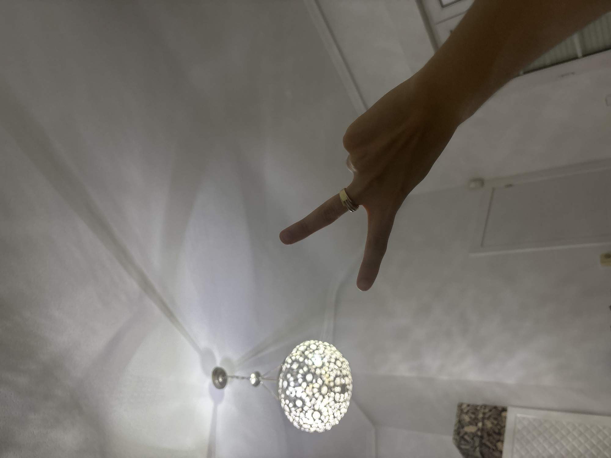 A hand with two fingers extended in a "V" shape, wearing a ring, is shown in the foreground. Behind the hand, a decorative ceiling light fixture hangs from the ceiling. The room appears to be a domestic interior, with a white ceiling and walls, and some furniture visible in the background. The lighting suggests it is nighttime or a dimly lit interior space, casting shadows on the ceiling. There is no information in the image to suggest the location is the city of Orihuela, Spain.