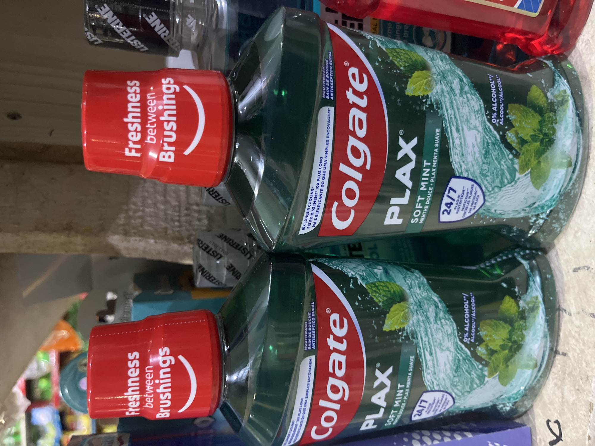 Two bottles of Colgate Plax Soft Mint mouthwash are displayed prominently in the foreground. Both bottles have red caps with white text and a white curved line resembling a smile. The bottles themselves are transparent green, containing green liquid and depicting mint leaves and water splashes. The labels on the bottles clearly show the "Colgate" logo and the "PLAX" brand name.
Behind the Colgate bottles, other products are visible on shelves, including what appears to be Listerine mouthwash bottles. The background suggests a retail environment, possibly a store or pharmacy. The overall lighting is somewhat dim, with some reflections on the bottles.
The scene is set in Bukuru, Nigeria, specifically within a retail establishment. There are no people visible in the frame. The objects are arranged on what appears to be a shelf or display area, indicating a commercial setting. The focus is on the Colgate mouthwash products.