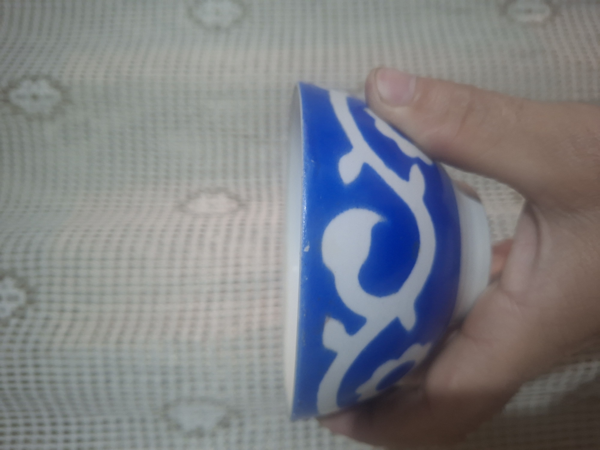A person's hand holds a small, blue and white patterned bowl in front of a light-colored, textured surface, potentially fabric or a table cloth. The scene's location is unknown.