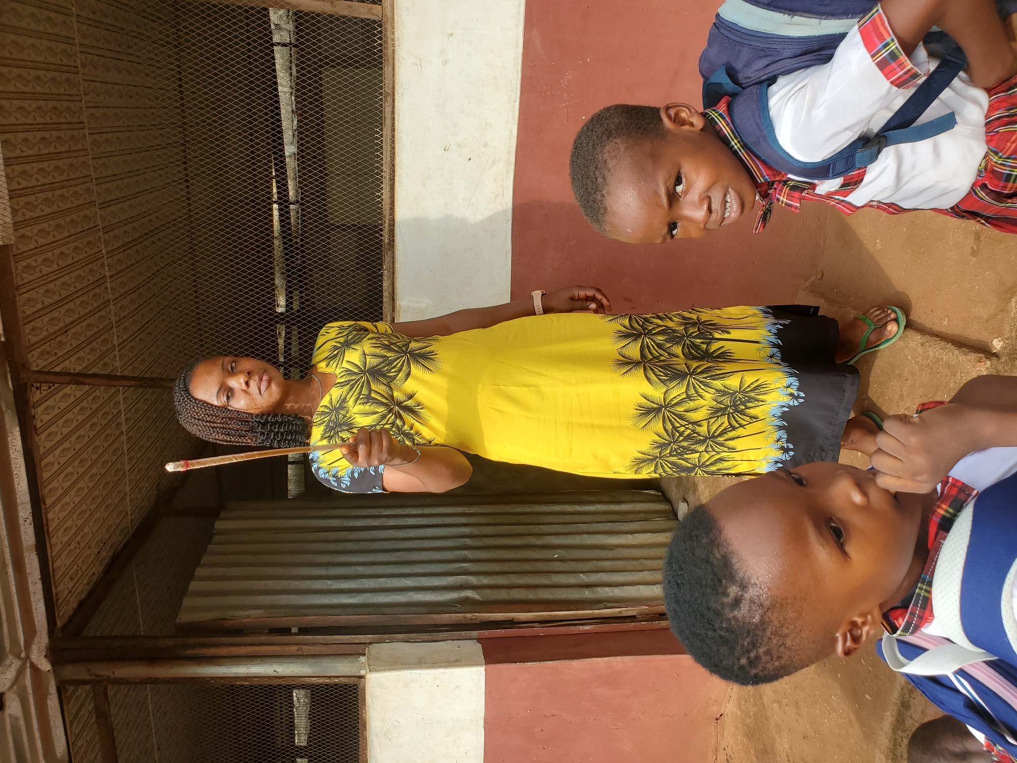 In Ukwu-Abwa, Nigeria, an adult woman with braided hair wears a yellow dress with black palm tree prints and black trim, holding a brown stick. Two children in school uniforms, including backpacks, are also present. The location appears to be outdoors, near a building with a metal screen and corrugated metal. The ground is reddish-brown. One child is looking at the camera, another child appears to be chewing. The woman has a watch on her left wrist.