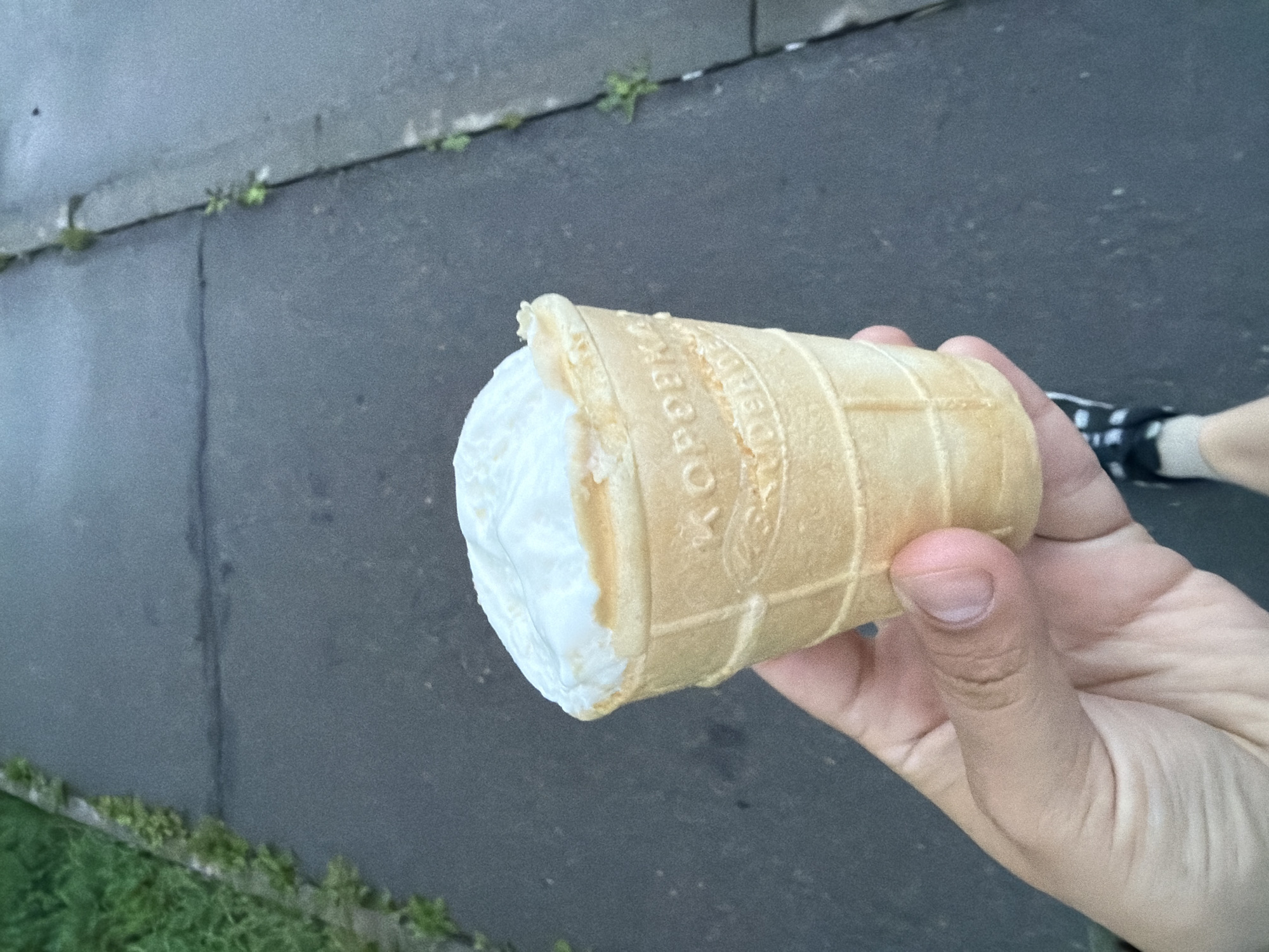 A person's hand holds a waffle cone filled with white ice cream against a blurred background of a paved walkway with grass on the left. The ice cream cone has text embossed on it that is partially visible, appearing to be "KOPOEEKA". The person's thumb and index finger are gripping the cone. The paved surface is dark gray and shows some cracks and debris. The grass on the left is green and appears to be growing along the edge of the walkway. The lower right portion of the image shows a glimpse of a person's leg wearing a black and white checkered shoe and a white sock. The scene is set in the city of Froly, Russia.