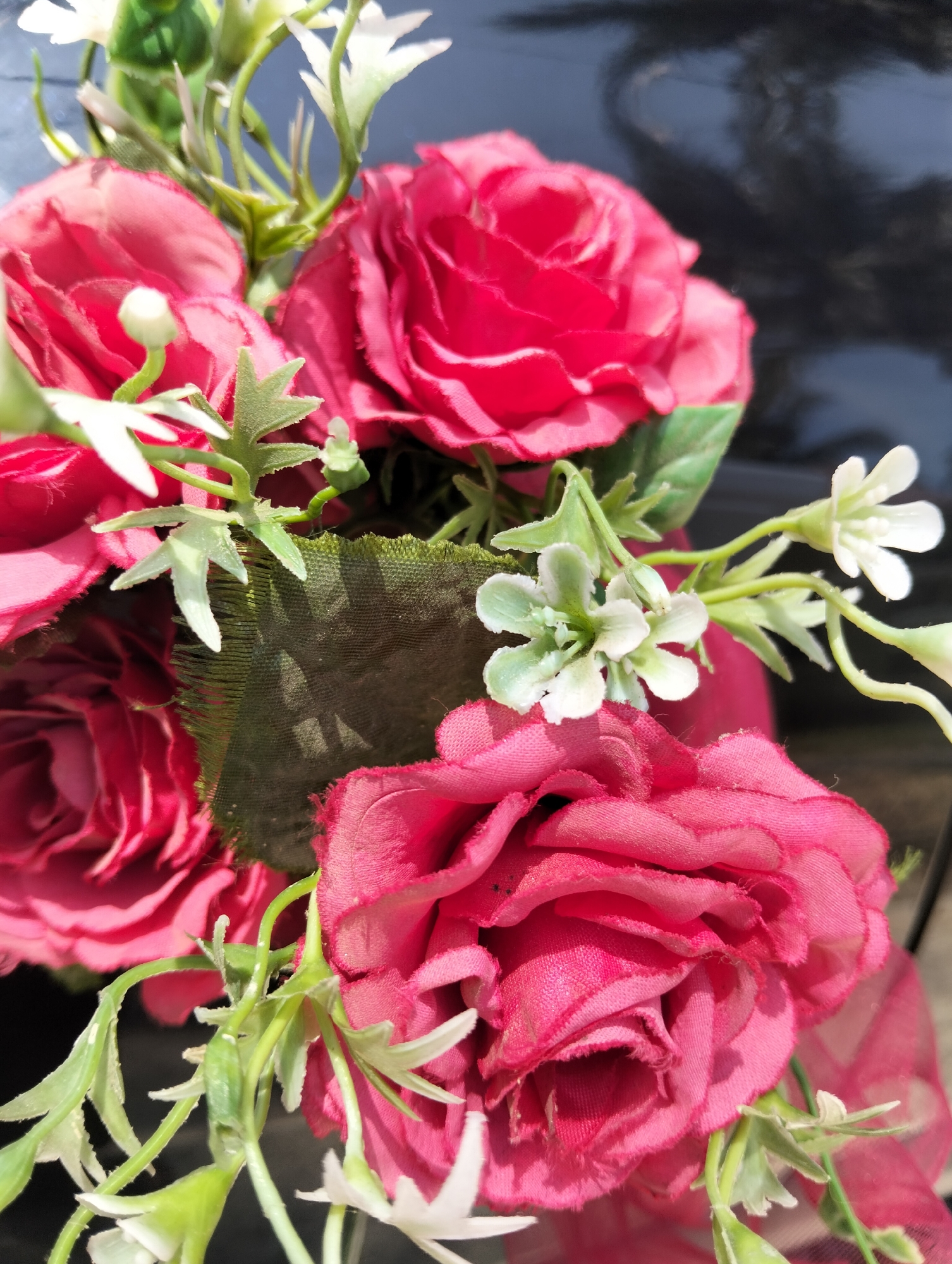 This is a close-up, eye-level shot of a bouquet of artificial pink roses. The roses are clustered together, with their vibrant pink petals taking center stage. Some of the roses are fully bloomed, revealing their layered petals, while others appear to be in bud form. Interspersed among the roses are artificial green leaves and delicate white flowers with multiple small petals. A red ribbon is visible in the lower right quadrant of the image, draped behind some of the flowers. The background is dark and reflective, possibly a polished surface, with subtle, indistinct shapes visible within it. The lighting suggests an outdoor setting, with sunlight creating shadows and highlights on the petals. The image does not contain any people or any identifiable elements of the city of Pulonga, Indonesia.