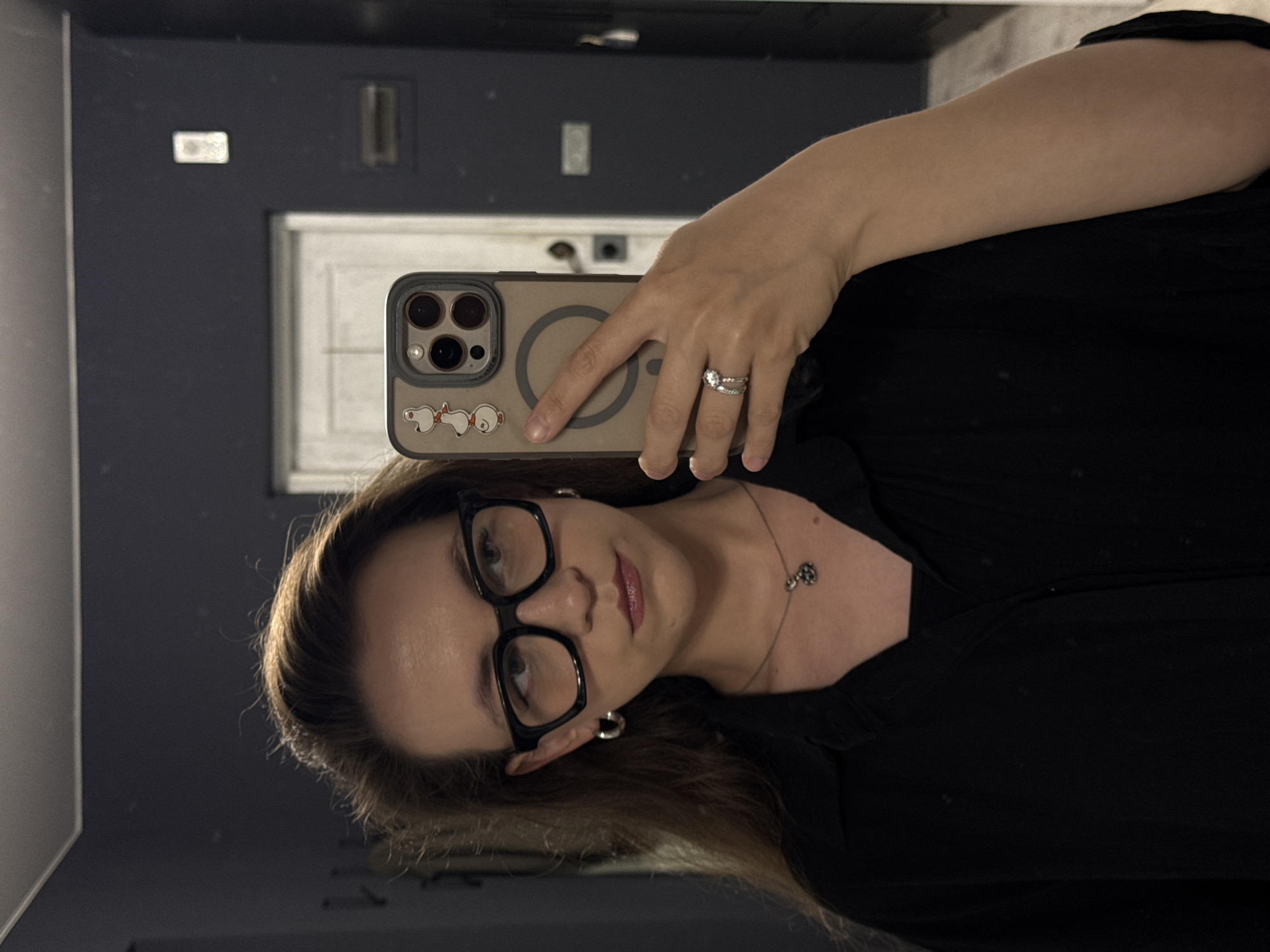 This is a vertical, slightly angled, mirror selfie of a woman wearing glasses and a black top. She is holding a smartphone in her right hand, which is adorned with two rings on her ring finger. The smartphone case features a cartoonish image of Snoopy and Woodstock. A delicate silver necklace with a small pendant is visible around her neck. The background, reflected in the mirror, shows a dark wall with a white door and what appear to be light fixtures. The lighting suggests it might be indoors, possibly in a home or public restroom. The overall mood is casual and posed.