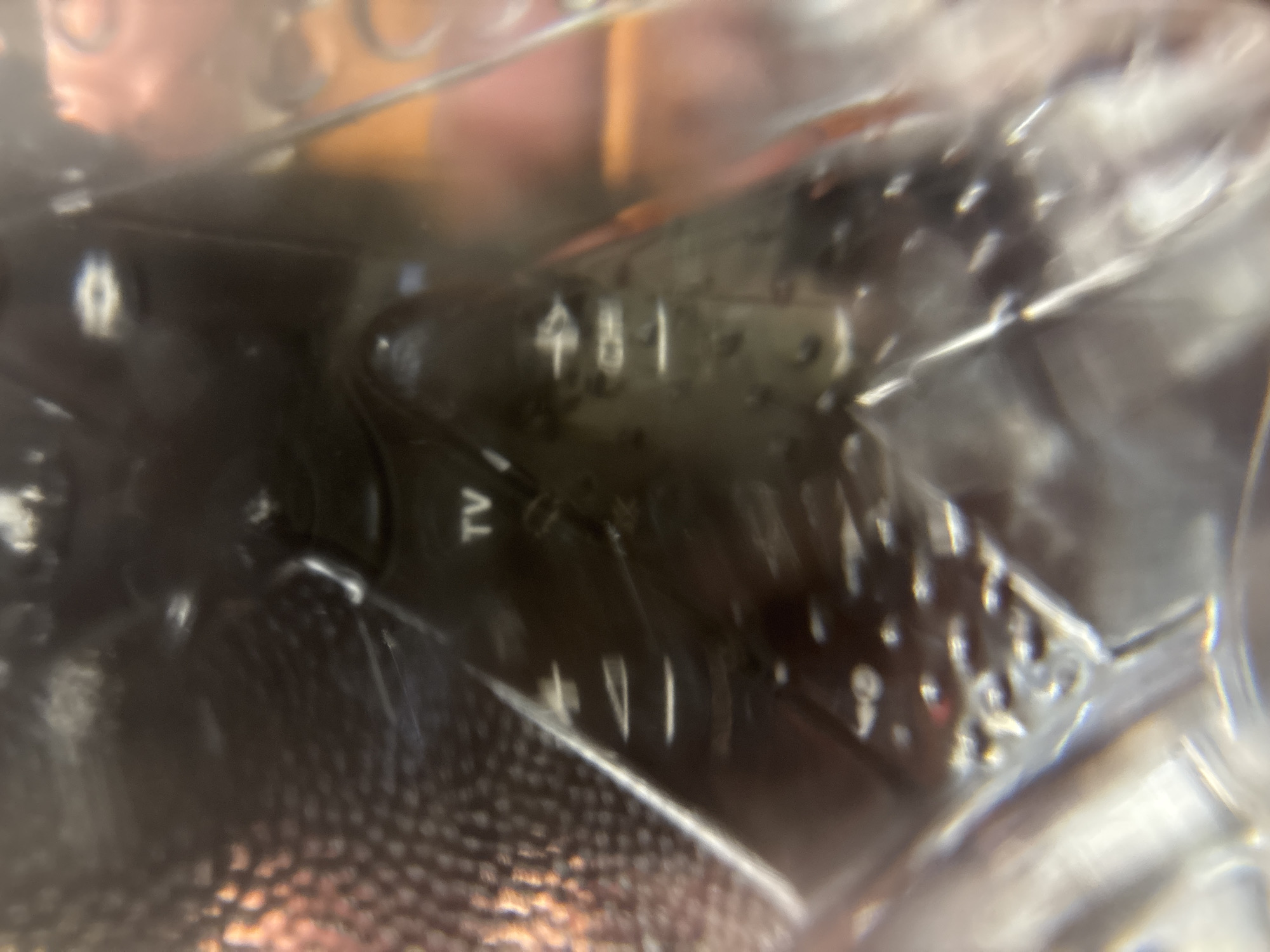 The image appears to be a close-up, out-of-focus shot of a remote control, likely for a television or audio device. The surface is black with some reflective qualities. Visible markings include the letters "TV" and what appear to be numerical indicators like "481" and another set of markings that are harder to discern due to the blurriness. There are also small, raised buttons or indicators visible. The lighting in the image is somewhat harsh, creating glare and making it difficult to identify finer details. The environment around the remote is not clearly defined but appears to be dark or dimly lit, with some brightly colored, out-of-focus elements in the background.