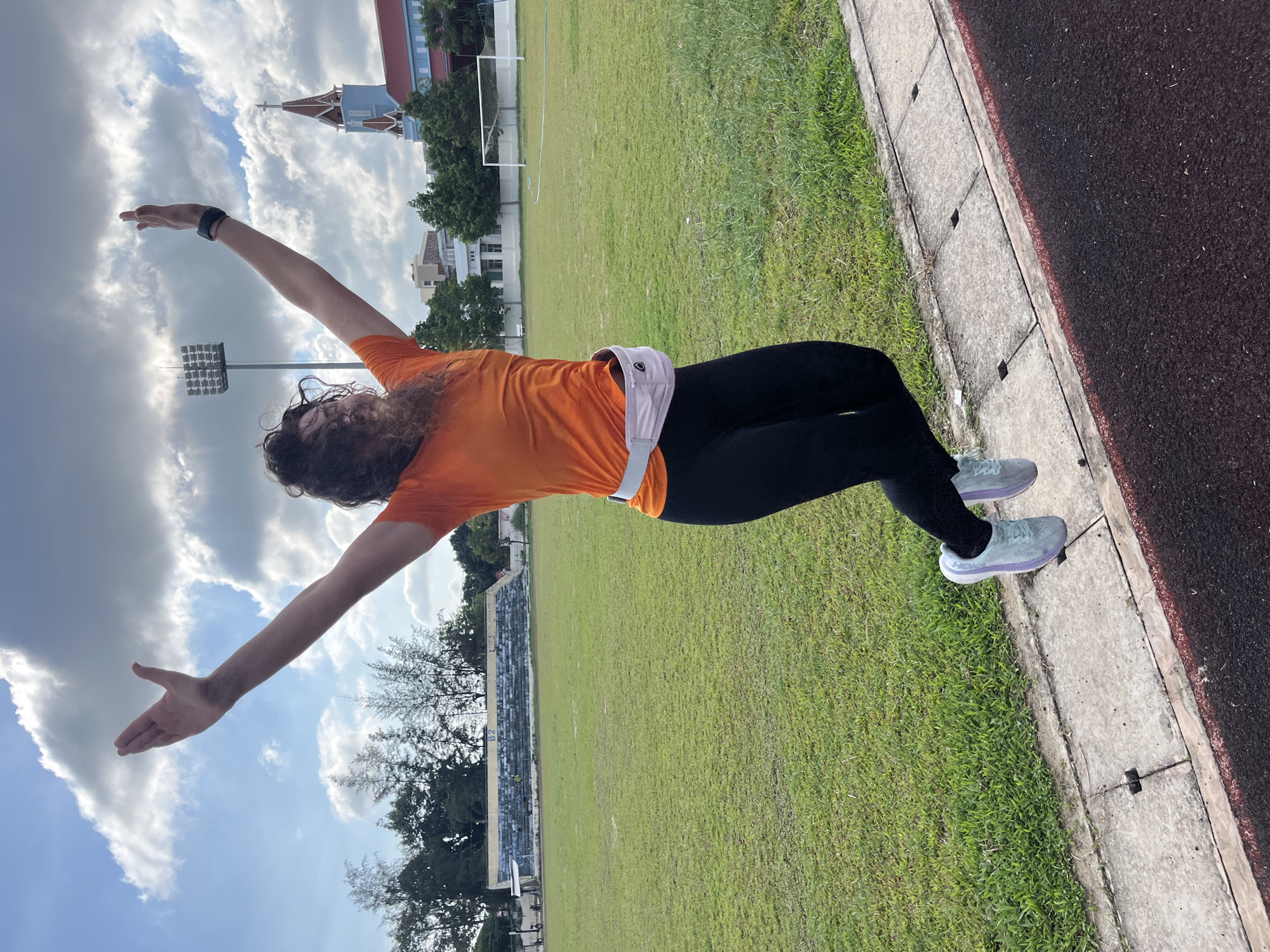 A woman with curly brown hair, wearing an orange t-shirt, black leggings, and light blue and purple athletic shoes, is captured mid-air in a joyful jump on an outdoor track. She has her arms raised and extended upwards, and a light pink fanny pack is visible around her waist. The background depicts a grassy field with a small set of bleachers and a soccer goal. In the distance, a prominent blue and red church with a tall steeple, a stadium light, and other buildings are visible under a partly cloudy sky. The scene is set in Phan Thiết, Vietnam.