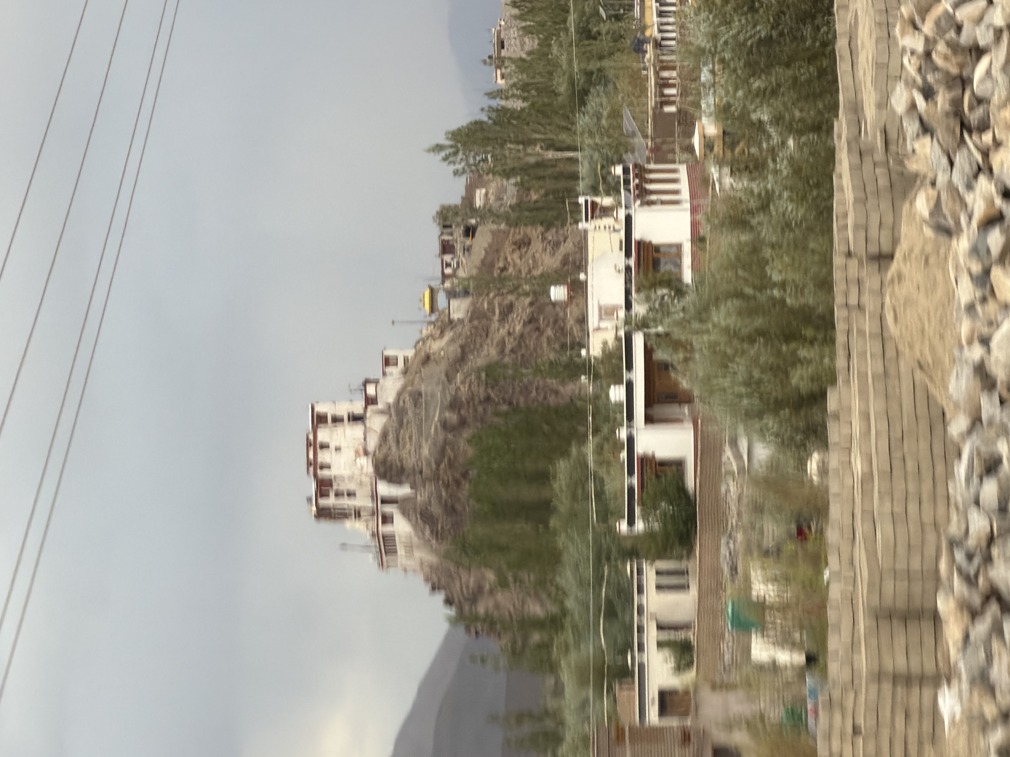 The image depicts a somewhat blurry, distant view of a white monastery complex situated on a rocky hillside under a pale, overcast sky. The monastery has multiple levels and windows, with some red trim visible. Below the monastery, a scattering of trees and some modern buildings with white walls and dark trim are visible. In the foreground, there are piles of bricks and rocks, and some greenery. The scene appears to be in Tanglakpurna, India, characterized by its mountainous terrain and unique architecture. There are no visible people or specific actions taking place.