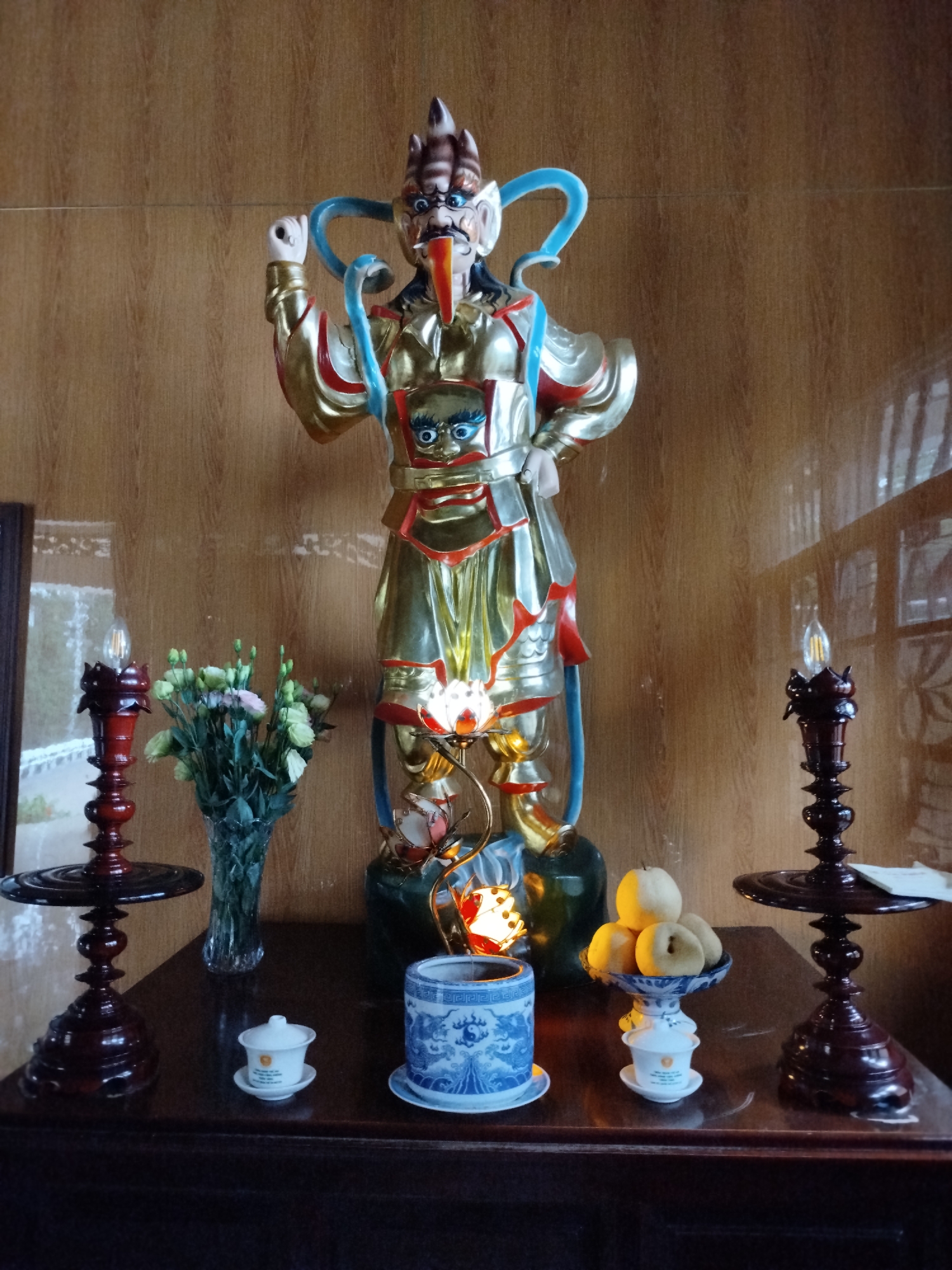 A statue of a deity stands on a dark wooden table in front of a wood-paneled wall. The statue is brightly colored, with gold, red, and blue accents. It has a distinctive face with a long, orange beak-like feature and a headdress with horn-like protrusions. Beneath the statue are several objects: a blue and white ceramic pot, a small white cup with a lid, and a dish holding several yellowish fruits. To either side of the statue are tall, dark wooden candleholders with electric candle bulbs. A vase of flowers is placed near the left candleholder. This scene is likely in Xuân Trường - Đà Lạt, Vietnam, based on the information provided.