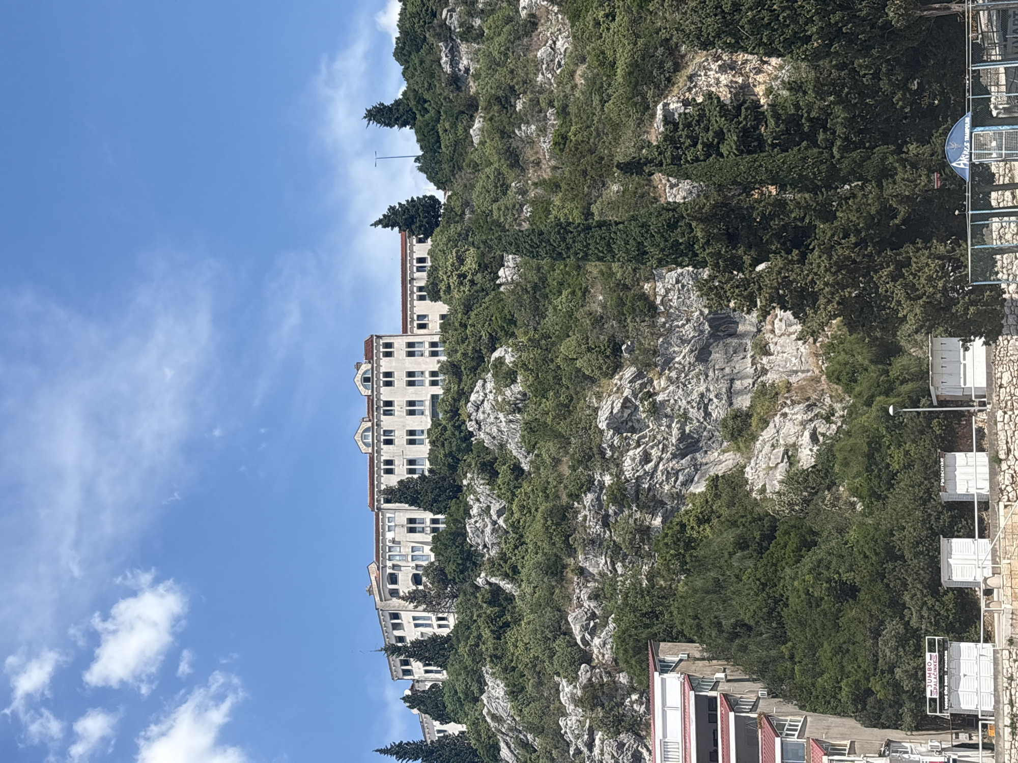 The image shows a hillside in Neum, Bosnia and Herzegovina. The hillside is covered in trees and rock outcroppings. A large building is situated on the hillside. The sky is blue with a few clouds. Some commercial signage is visible on the right side of the image.
