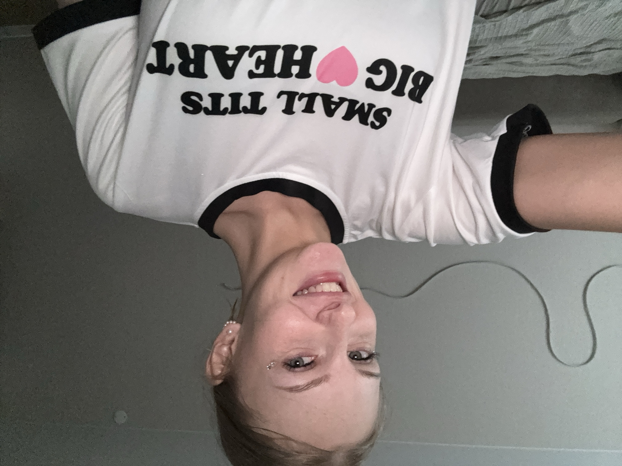 The image shows a young woman with blonde hair and blue eyes, smiling at the camera. She is wearing a white t-shirt with black trim and a graphic that reads "SMALL TITS BIG HEART" with a pink heart symbol. She has piercings on her face and ears. The background appears to be a room with a wall and a bed. There are no discernible features that would indicate the location is the city of Perm, Russia.