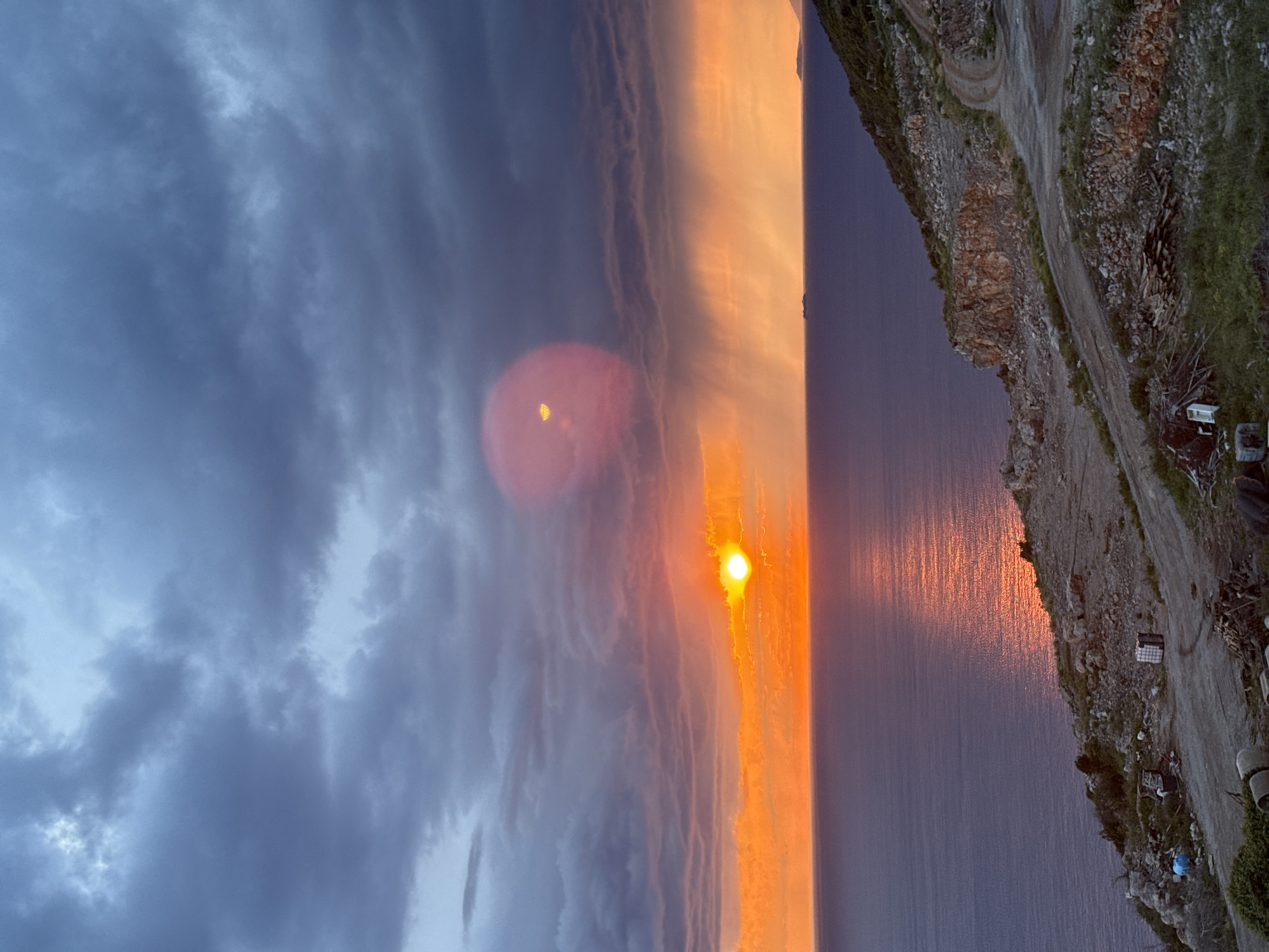 The image shows a sunset over water next to a rocky cliff in Kovačko Polje, Montenegro. The sky transitions from dark blue with storm clouds to orange-yellow light around the sun. The water surface reflects the golden sunset. A road and some vegetation are visible on the cliff top. There are some structures, like containers, near the road.