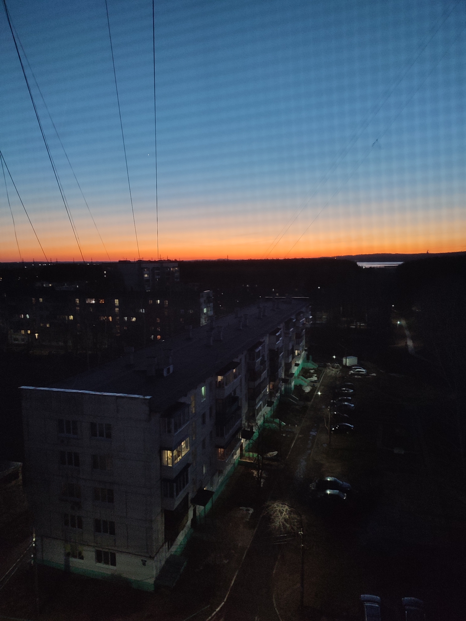The image depicts a view from a high vantage point in the city of Chaikovsky, Russia, likely from an upper floor of an apartment building. The scene is at twilight or dusk, with a gradient sky transitioning from orange at the horizon to deep blue overhead. Overhead power lines are visible, obscuring the sky. Below the horizon, a residential area is visible, with several multi-story apartment buildings, mostly shrouded in darkness. Some windows are illuminated. A street is visible alongside one building, with parked cars and streetlights casting faint illumination. The overall environment appears urban and residential.