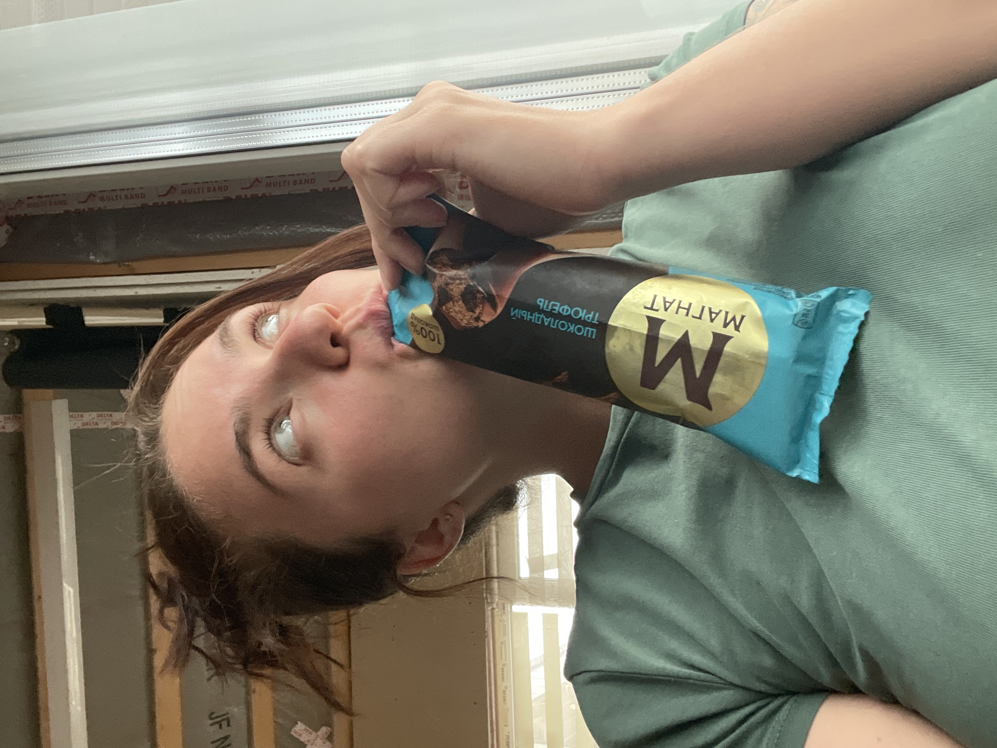 A woman is depicted in an indoor setting, presumably in Мосметаллоконструкция Онт, Russia, as text in the image indicates Russian writing on the packaging of what appears to be a chocolate ice cream bar. She is holding the ice cream close to her face as if she is about to take a bite. The packaging is blue and brown with the brand name "МАГНАТ" displayed prominently. The woman is wearing a green t-shirt. In the background, there is a window with a partially visible frame. The photograph is taken at an angle, making the scene appear slightly tilted.