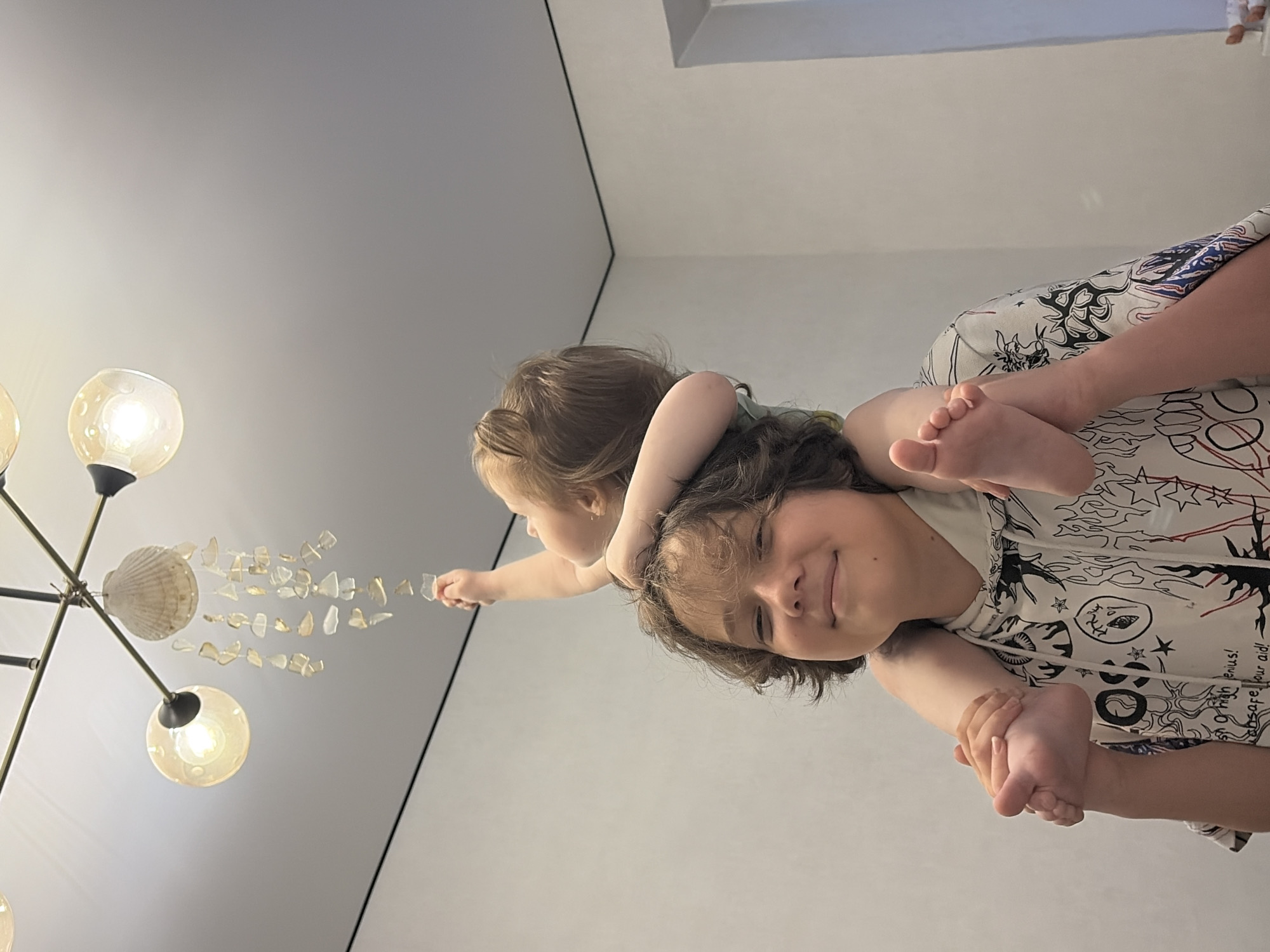 Two children are depicted in the image, with the older child carrying the younger child on his shoulders. The younger child, a toddler, is reaching up towards a chandelier. The older child, a pre-teen, is smiling and looking forward. The chandelier is a decorative piece hanging from the ceiling, featuring a shell at the top and various glass fragments suspended below. The setting appears to be an indoor environment with a white ceiling and walls. No specific details about the city of Chaikovsky, Russia, are present in the image.