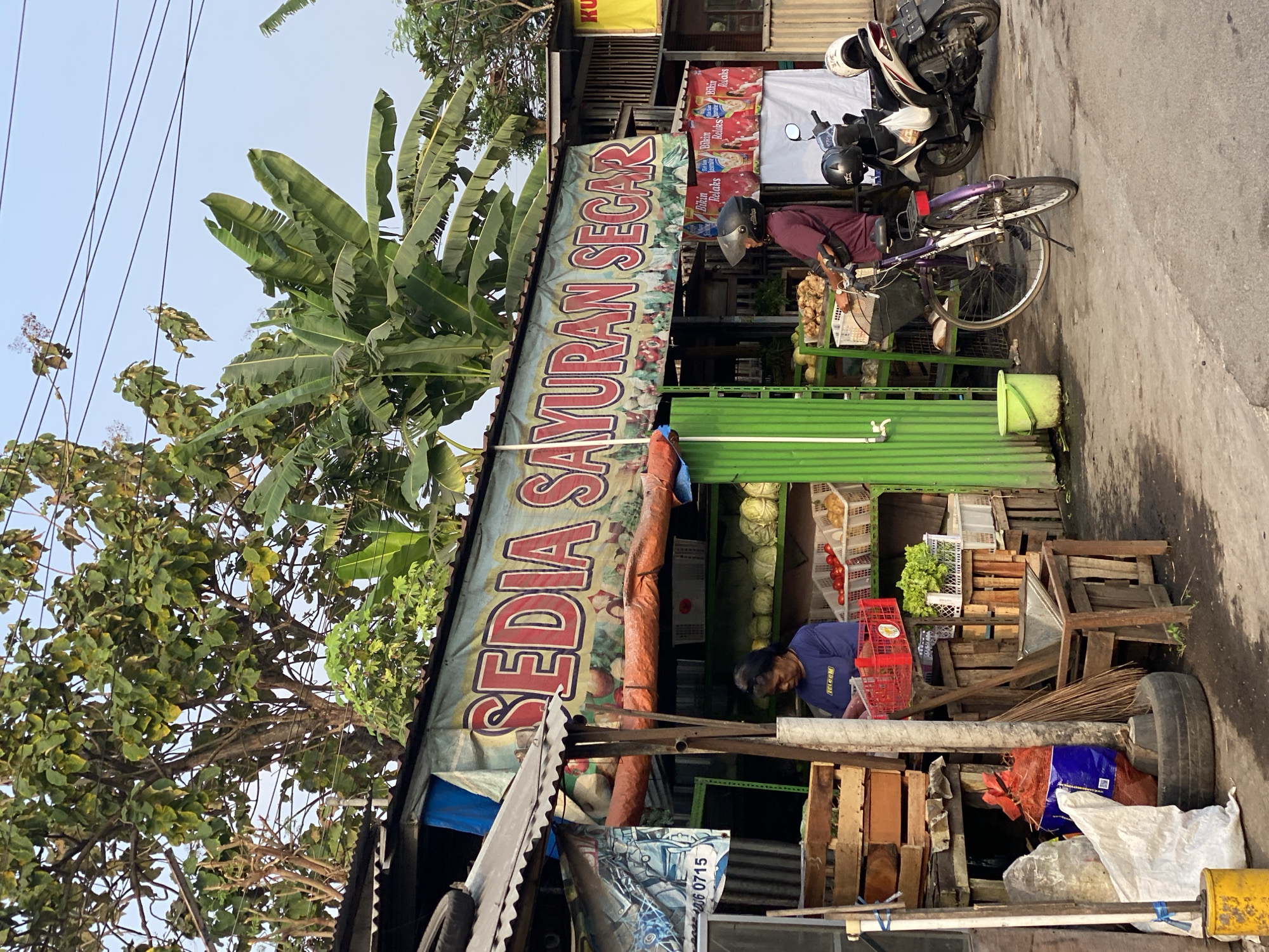 In Kasihan, Indonesia, a roadside vegetable stall displays a banner reading "SEDIA SAYURAN SEGAR," meaning "Fresh Vegetables Available." A woman in a blue t-shirt stands behind the counter, arranging produce. A person wearing a helmet is on a bicycle to the right of the stall, and a motorcycle is parked nearby. The stall is stocked with various fresh vegetables, including cabbage, tomatoes, and leafy greens, arranged in baskets and crates. Wooden structures and corrugated metal roofing form the stall's framework. The scene is set against a backdrop of lush green trees and a clear sky, with overhead power lines indicating an urban or semi-urban environment.