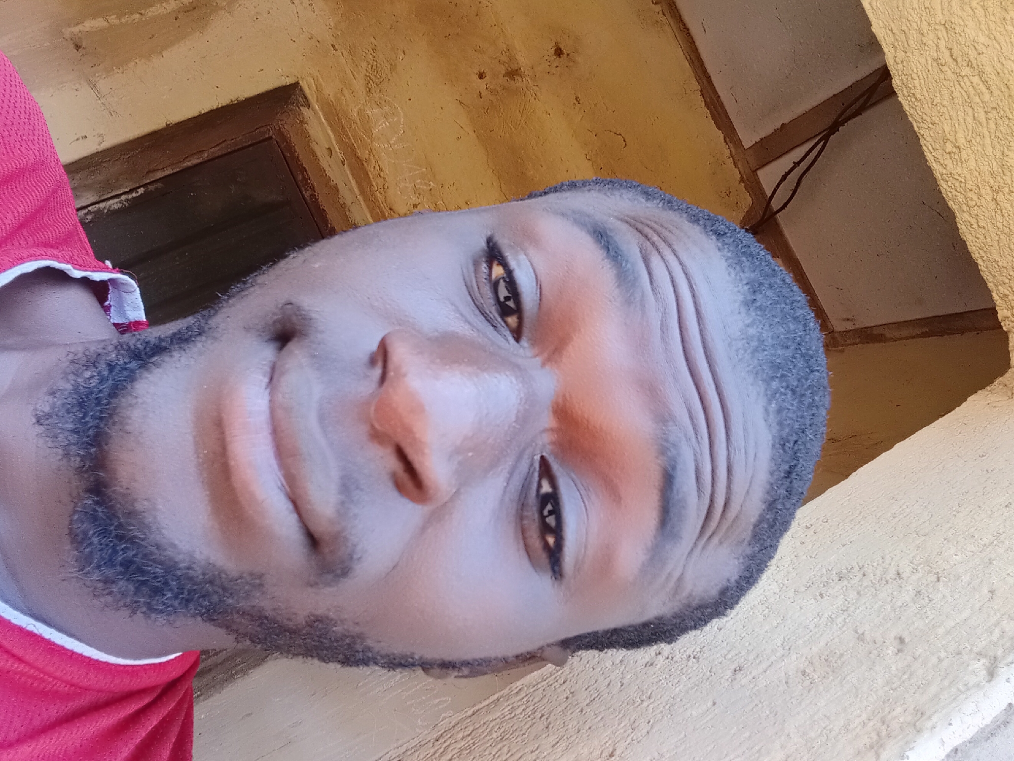 The image is a close-up, selfie-style photograph of a Black male in Bukuru, Nigeria. He is wearing a red t-shirt with white trim around the collar. His facial features are visible, including his eyes, nose, mouth, and facial hair. The background appears to be an outdoor environment with textured walls and some exposed wooden beams and wires, suggesting an unfinished or rustic structure. The lighting indicates it is daytime.