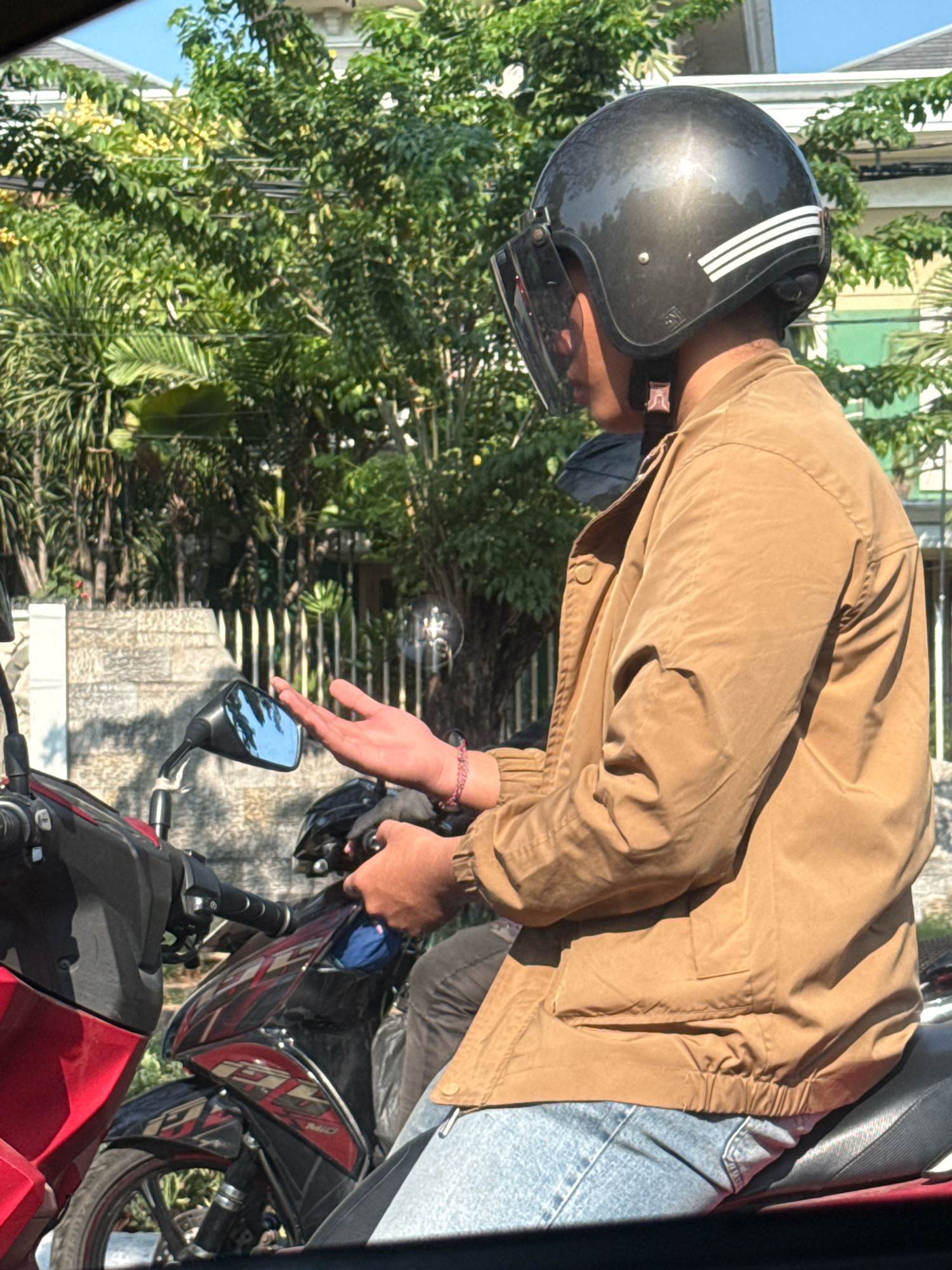 In Surabaya, Indonesia, a person wearing a brown jacket, blue jeans, and a black helmet with a clear visor is seated on a red and black motorcycle. The person's left hand is raised, palm facing outward, and they are wearing a thin pink bracelet. The motorcycle is parked outdoors, with lush green foliage and a building visible in the background under a clear blue sky. Sunlight casts shadows on the scene.