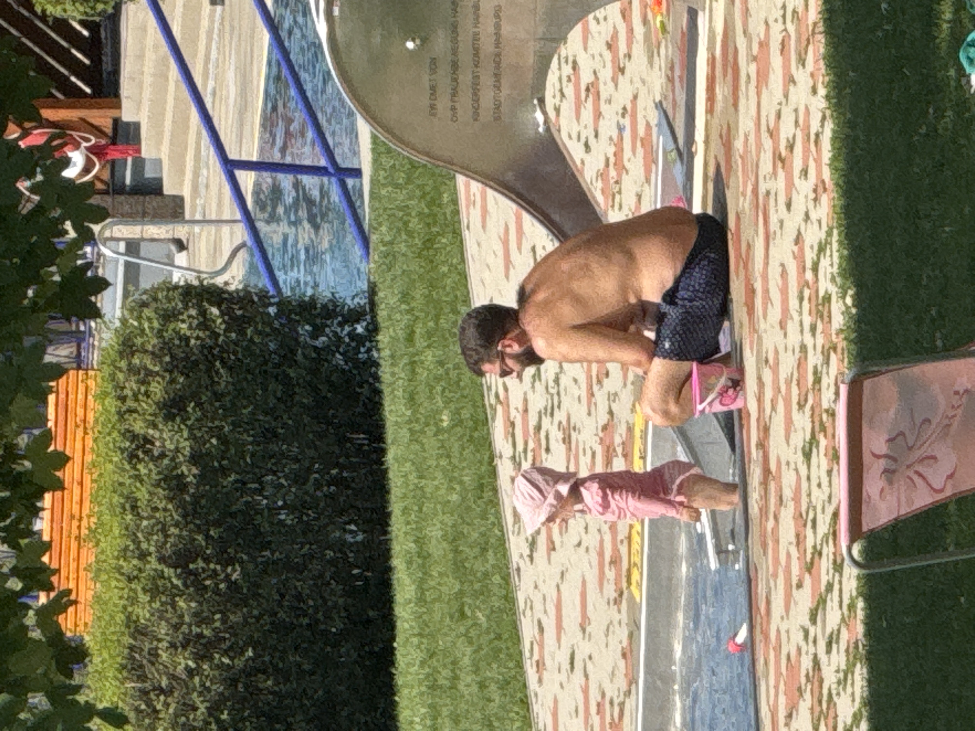 The image depicts a sunny outdoor scene at a public swimming pool in Hainburg an der Donau, Austria. A man with dark hair and sunglasses is sitting shirtless, wearing dark patterned swim trunks. He is looking towards the left. To his left and slightly in front of him, a young child wearing a pink sun hat and a pink swimsuit stands with their back to the camera, facing the water. The child is looking down towards a pink bucket on the ground. In the background, a blue railing leads into a swimming pool. Further back, there is a large green hedge, a wooden structure with steps, and part of a waterslide. The ground around the pool is paved with light-colored tiles with an orange and green patterned border. A pink lounge chair is visible in the foreground, on a patch of green grass.