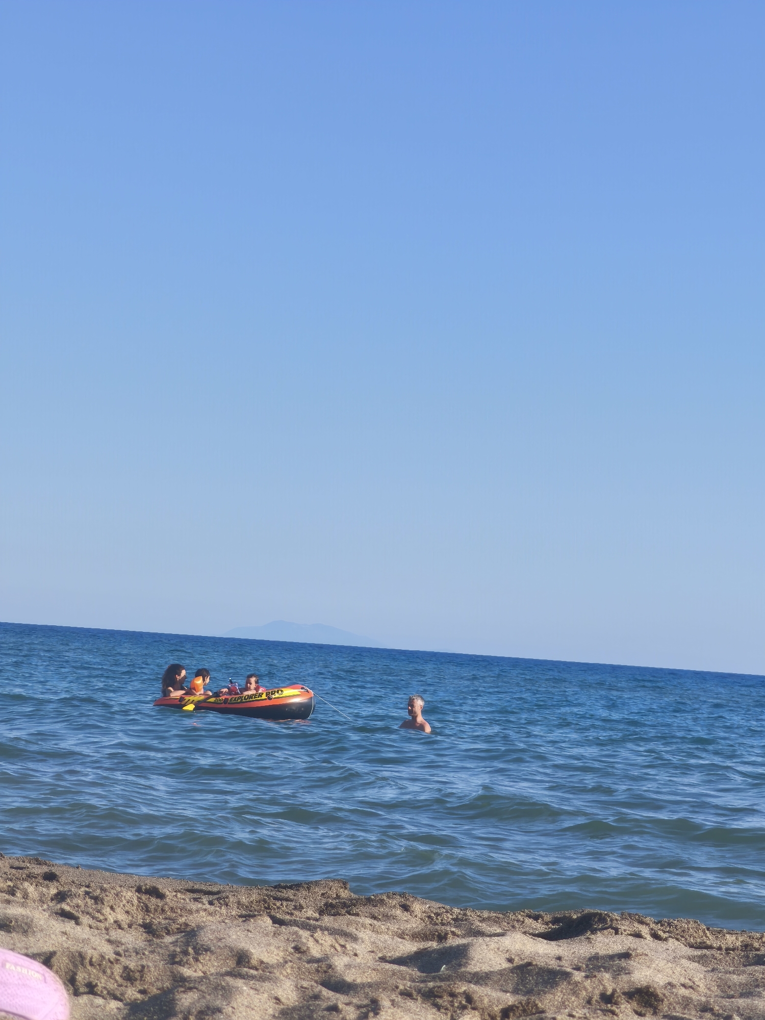 In the Tyrrhenian Sea off the coast of Castiglione della Pescaia, Italy, on a clear, sunny day, a bright orange and yellow inflatable tube is being towed by an unseen boat. Four individuals, appearing to be a family, are riding on the tube. A man with short, dark hair is in the front, steering or holding on. Behind him, a woman with dark hair is seated, also holding on. Two children are positioned further back on the tube. To the right of the inflatable tube, a man with light-colored hair is swimming in the sea, facing away from the camera. The water is a vibrant blue, with small waves rolling towards the sandy beach in the foreground. The beach is covered in fine, light-brown sand, with some debris and footprints visible. In the distance, a faint, hazy outline of a mountainous island can be seen on the horizon under a clear, pale blue sky. The overall scene depicts a leisurely day at the beach, with water sports and swimming.