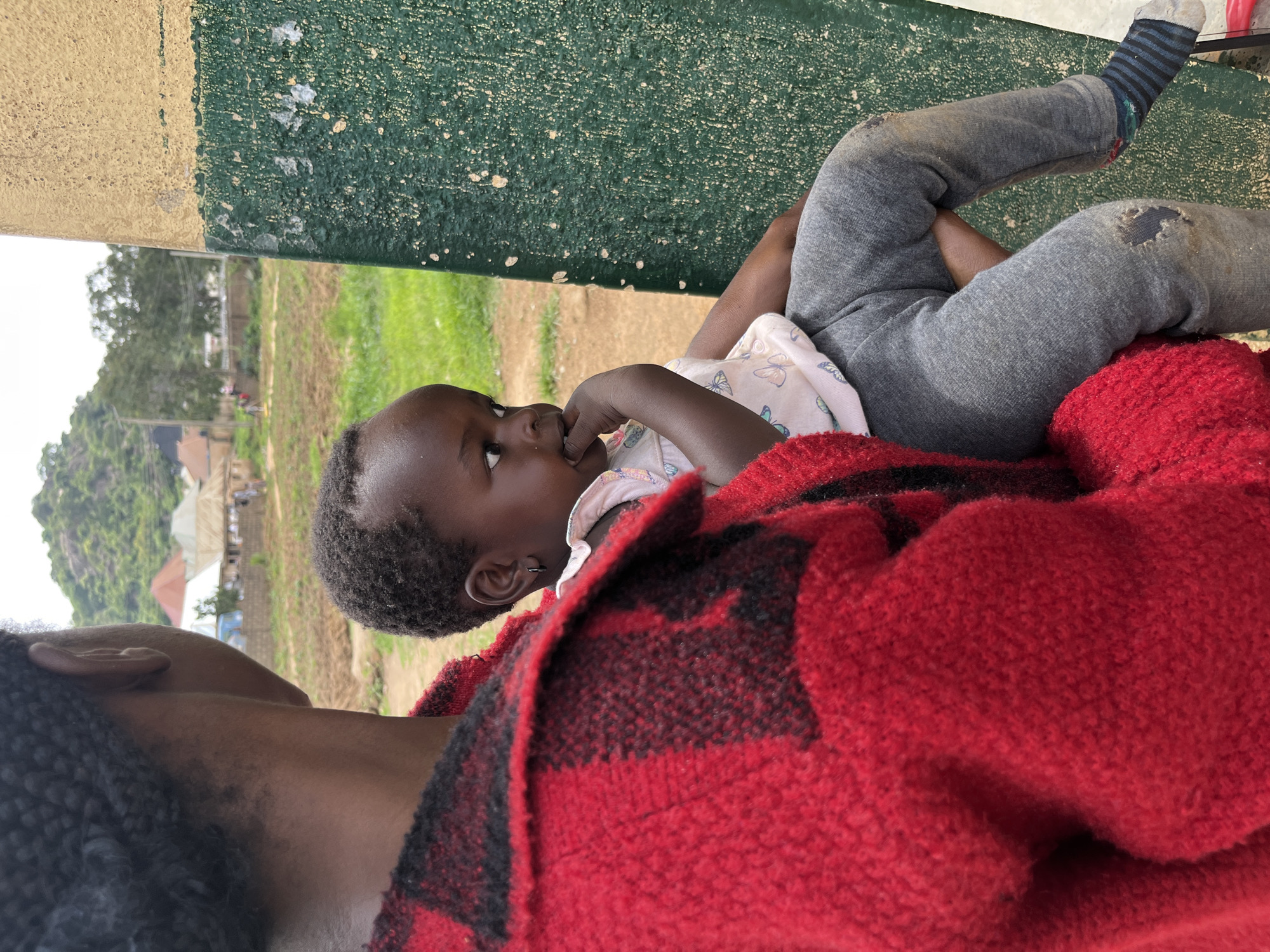 In Bukuru, Nigeria, a close-up shot focuses on a young child being held by a person whose back is to the camera. The child, facing left, has dark hair and is wearing a light-colored shirt with a butterfly pattern and grey pants. The child's right hand is near their mouth. The person holding the child is wearing a red and black plaid jacket. The background shows a slightly blurred view of a village with buildings and a green, textured wall to the right.