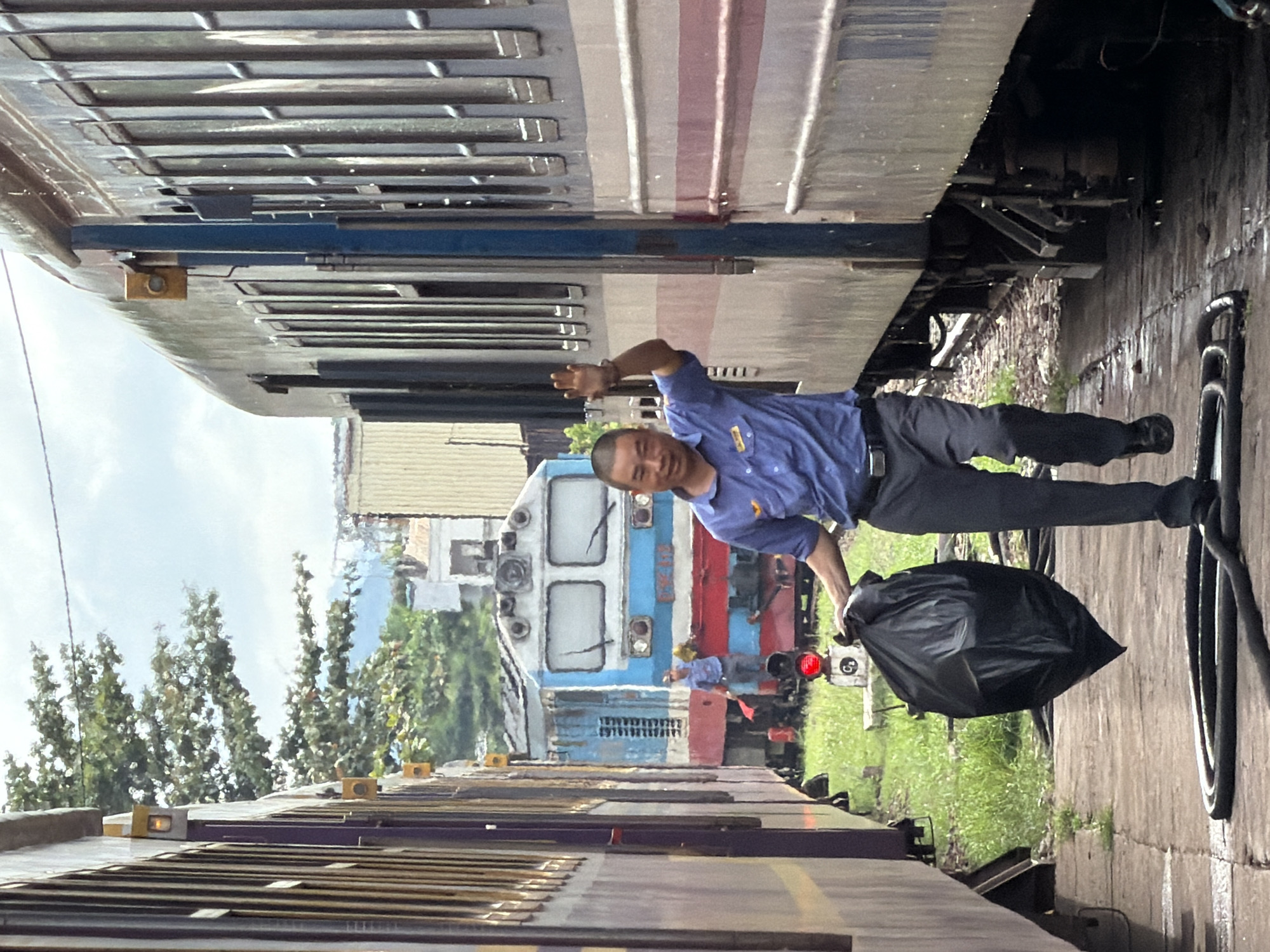 An overcast day at a train yard in Da Nang, Vietnam, features two trains parked on parallel tracks. In the foreground, a man in a blue uniform carrying a large black trash bag walks towards the viewer, waving with his right hand. He wears a dark belt and dark trousers, and black shoes. To his left, a train with purple and yellow accents is visible. To his right, a train with a light blue and red body is parked, and another man in a blue uniform is seen in the distance holding a red signal flag. The ground is a mixture of concrete and gravel, with lush green grass growing between the tracks. The sky is a pale blue with scattered clouds.