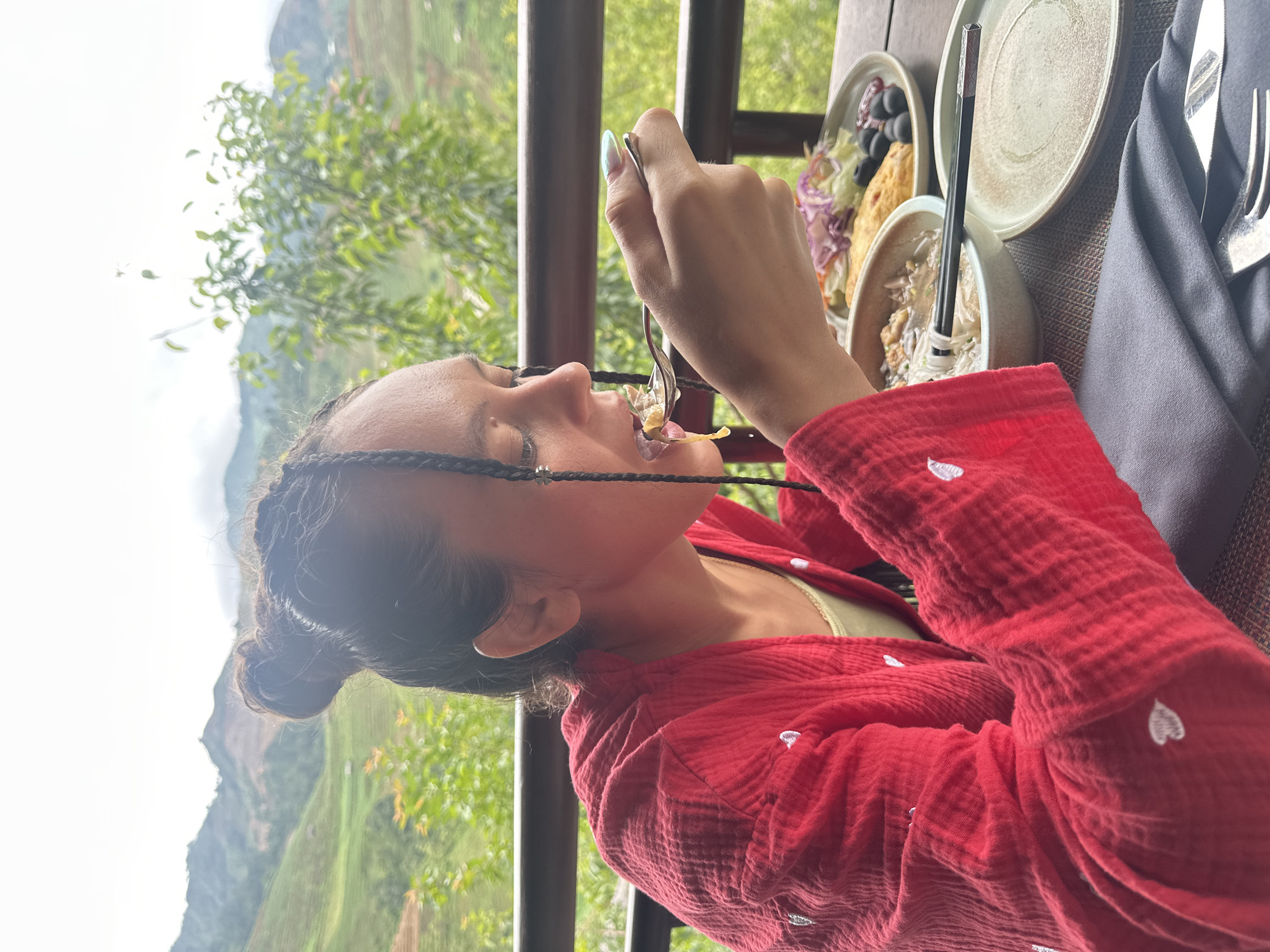 An adult woman is depicted in a side profile, seated outdoors on what appears to be a balcony or terrace, in Púng Luông, Vietnam. She is positioned to the left of the frame, facing right, with her eyes downcast towards a spoonful of food she is bringing to her mouth. Her dark hair is styled in a high bun, with several thin braids framing her face, some adorned with small silver, star-shaped embellishments. She wears a red, textured, long-sleeved garment featuring embroidered white heart motifs, over a light yellow-green undergarment. Her right hand, with long, pointed nails painted with a greenish-blue iridescent polish, holds a silver spoon from which she is consuming a portion of food.
On a dark, woven table in front of her, a meal is laid out. This includes a light-colored bowl containing noodles or soup with dark chopsticks resting on its rim, and a grey/beige plate holding a salad composed of purple cabbage, lettuce, and red onions, alongside what appears to be an omelet or frittata and several dark olives or berries. An additional empty grey/beige plate is also present. A dark grey folded fabric napkin, holding a silver fork and knife, is situated in the foreground.
The background reveals a panoramic view of rolling green mountains, consistent with terraced agricultural landscapes common in Púng Luông. Lush green foliage and trees are visible immediately beyond a dark metal railing, which delineates the edge of the terrace. The sky overhead is bright and appears overcast.
