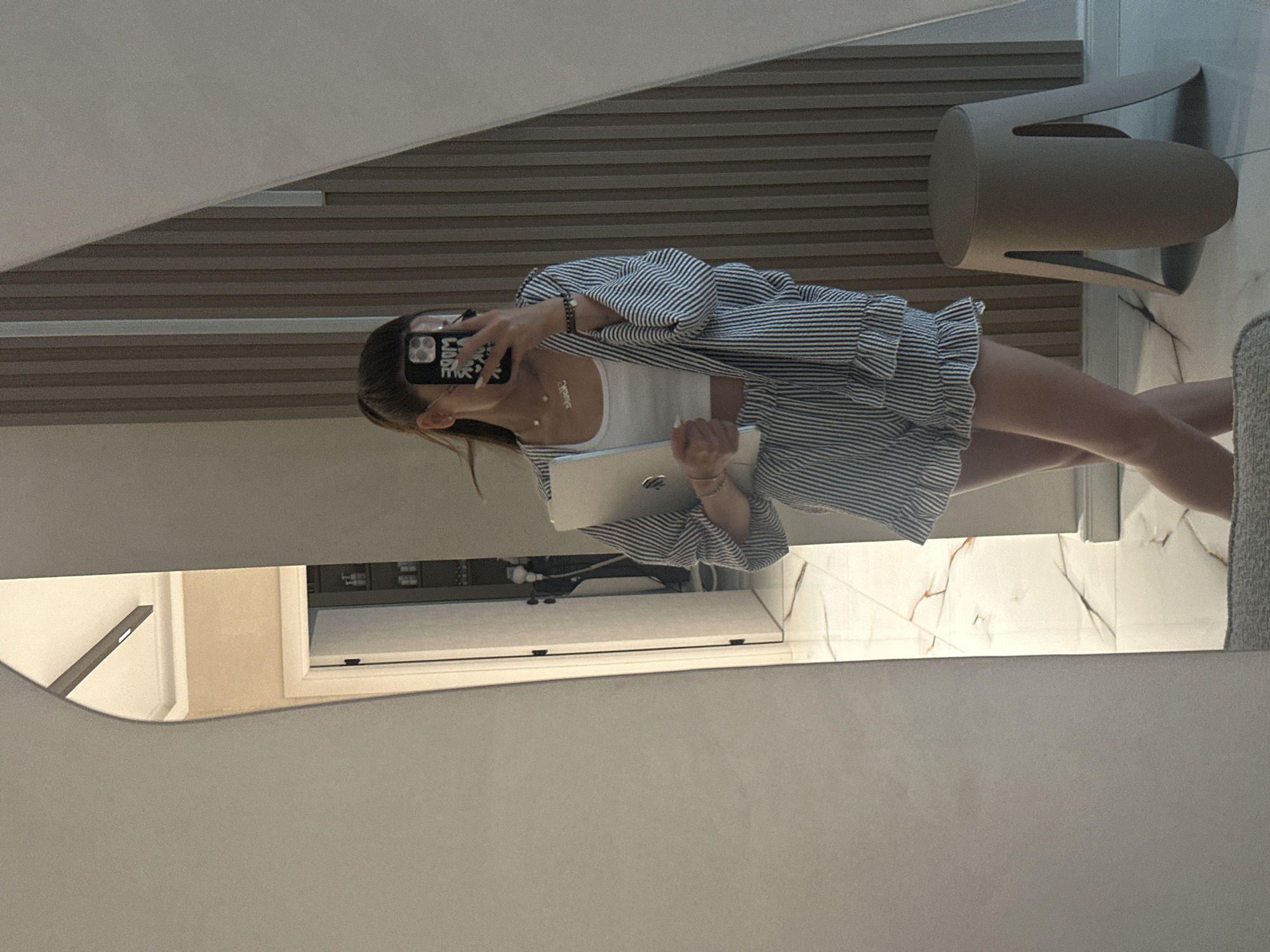 A young woman is reflected in a mirror in an indoor space, possibly a home in Petrich, Bulgaria. She is wearing a white crop top and matching striped shorts and a matching striped open robe. She holds a silver laptop in her right arm. She is also holding a black smartphone in her left hand, taking the photo. The background behind her in the reflection shows a wall with vertical wooden slats and a door. To the right of the woman, a taupe-colored stool is visible. The floor in the reflection appears to be tiled with a marble pattern. The mirror itself is large and has an irregular, curved edge on the left side.