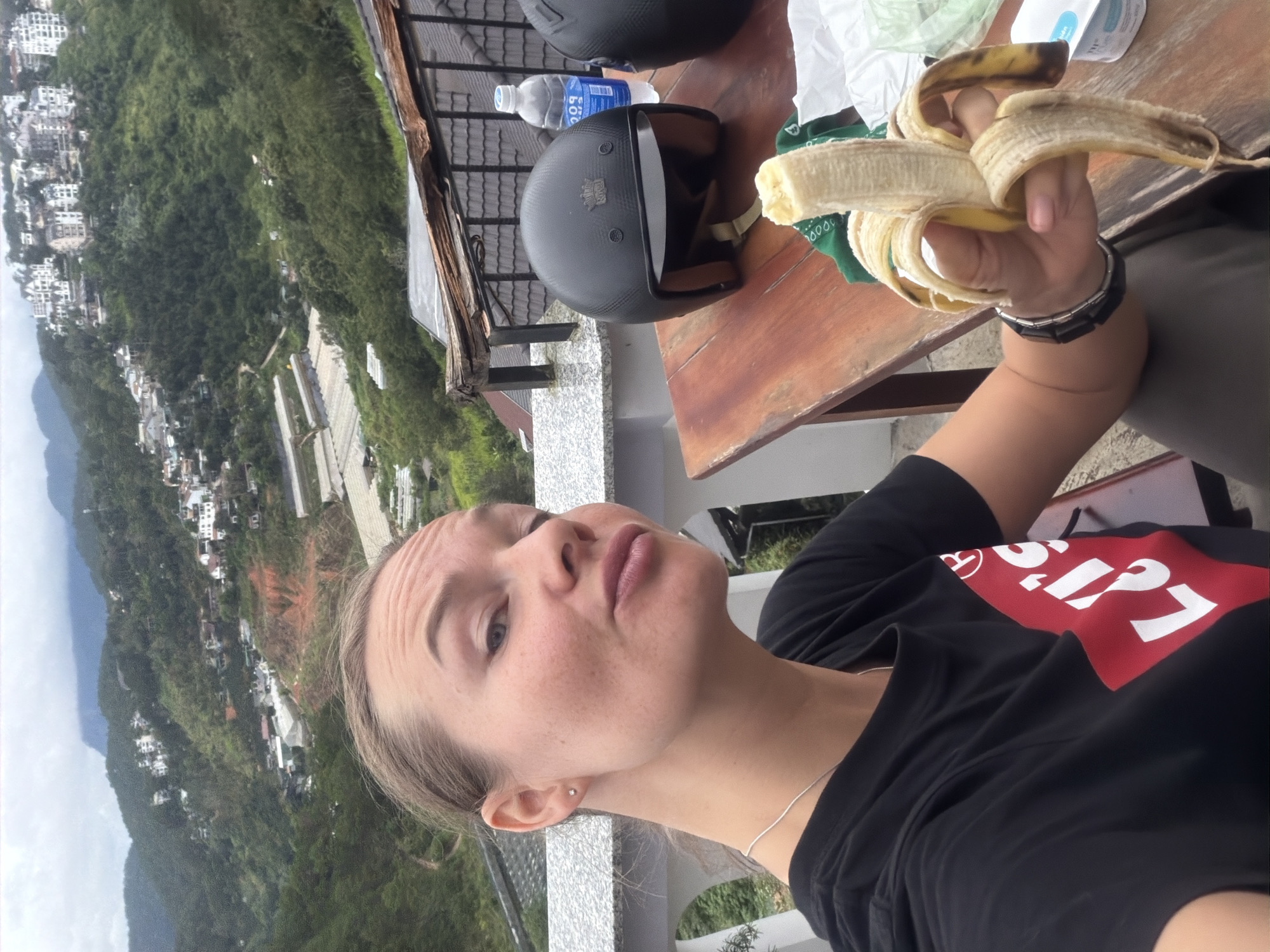 A woman with blonde hair pulled back is taking a selfie while holding a partially peeled banana. She is wearing a black t-shirt with a red and white Levi's logo. Her left ear has a small stud earring, and she is wearing a silver necklace. She has a bracelet on her left wrist. In the background, a mountainous landscape is visible, dotted with houses and buildings, characteristic of the hilly terrain of Da Lat, Vietnam. To the right of the woman, on a wooden table, there is a black motorcycle helmet, a bottle of water, and another helmet. The overall scene suggests an outdoor setting, possibly a viewpoint or a cafe in Da Lat.