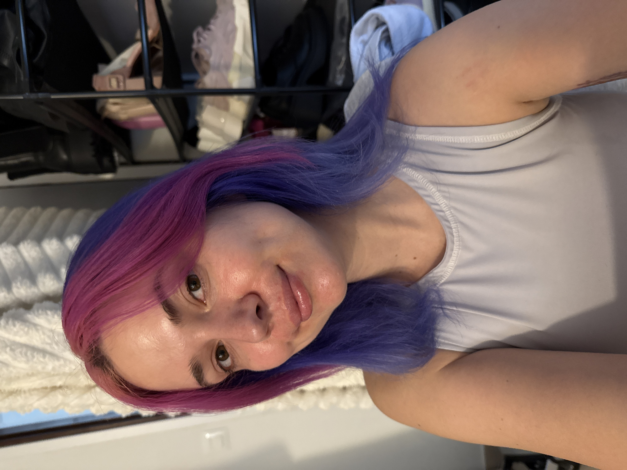 The image presents a close-up portrait of a young adult female with striking two-toned hair, featuring vibrant pink/magenta at the roots and mid-lengths, transitioning into a distinct purple/lavender hue towards the ends. She looks directly at the camera with a calm expression and a subtle, natural smile. Her eyes are brown, and her lips appear glossy. She is wearing a light grey or white athletic-style tank top.
The setting is indoors, likely a personal living space such as a bedroom or dressing room, located in the City of Zagreb, Croatia. The background is dimly lit and out of focus, revealing a multi-tiered shelf or wardrobe unit containing various items like shoes, bags, and clothing, suggesting a casual and lived-in environment. To the lower left of the frame, a textured white fabric, possibly a blanket or a rug, is visible, with a hint of a window or reflective surface beneath it. The lighting suggests it is daytime. The image captures a candid moment, possibly a selfie or a casual portrait. There is no visible text in the image.