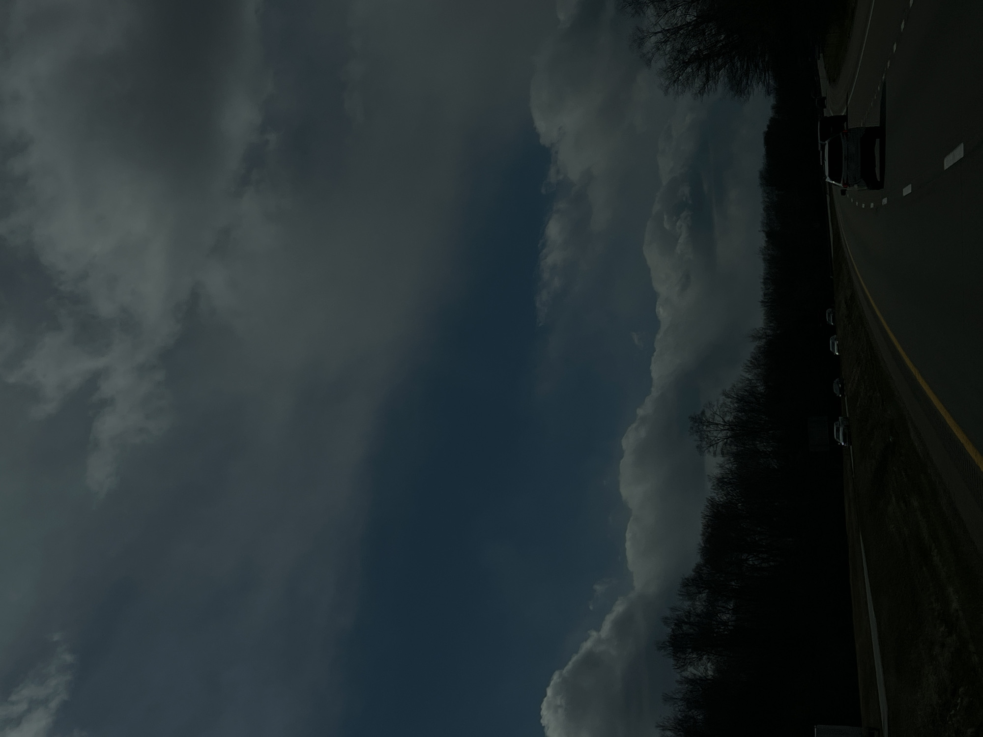 A dimly lit photograph shows a highway running to the right of the frame. A vehicle is present on the road. Alongside the highway, a dark, shadowed area filled with trees and greenery is visible. The sky above is filled with clouds, some dark and overcast, adding to the somber atmosphere of the image. This photograph was taken near the Town of Fishkill, United States of America.